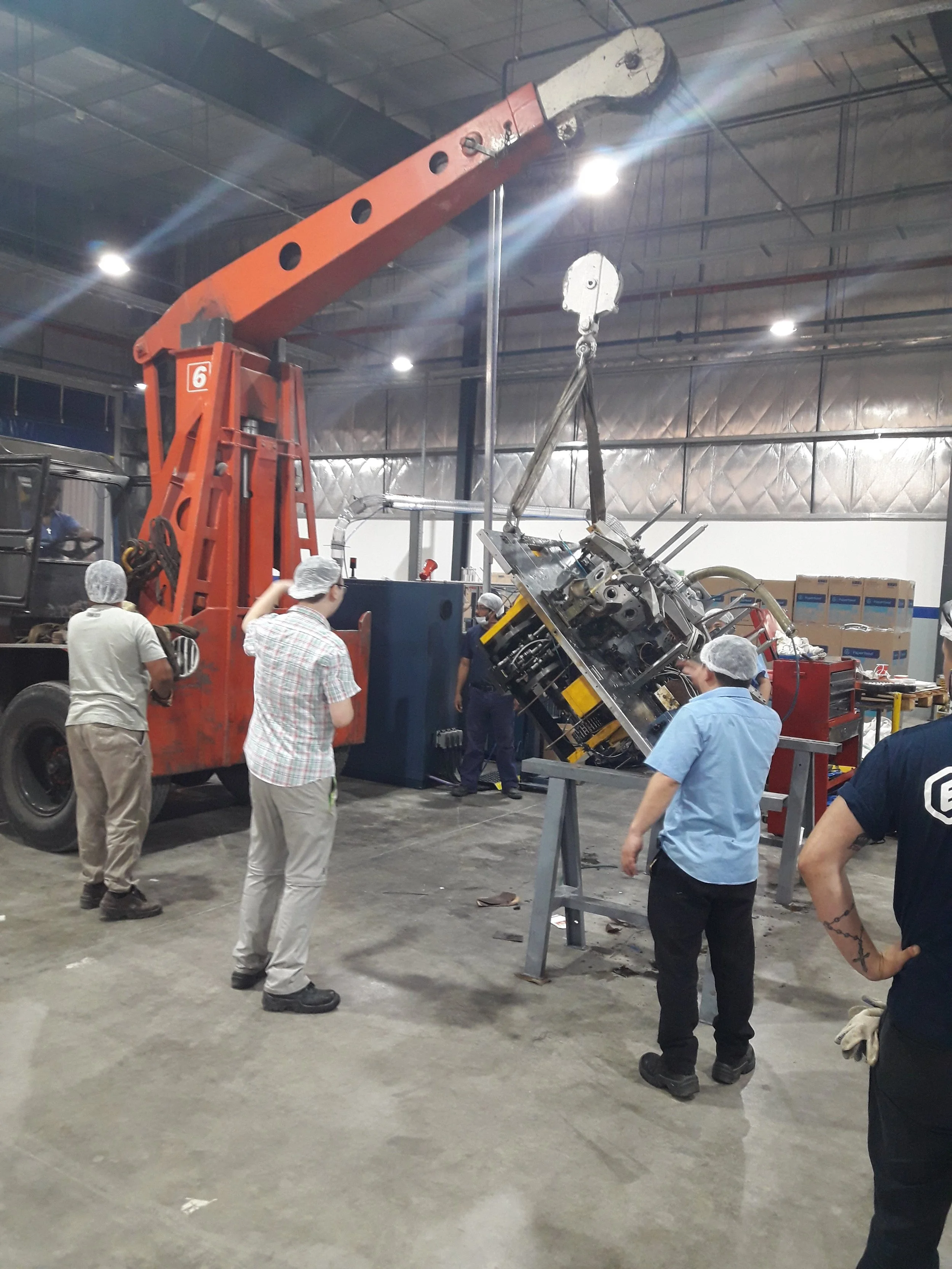 Varios empleados en un taller de reparación de maquinaria, observando la reparación de una pieza industrial grande, que está suspendida por un grúa dentro de un espacio cerrado.