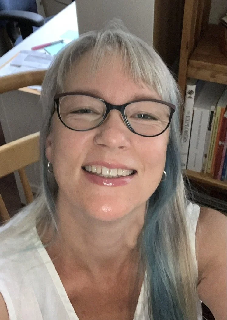 Close-up photo of a smiling woman wearing glasses, with blue streaks in her blonde hair, in a home setting with bookshelves and a desk in the background.