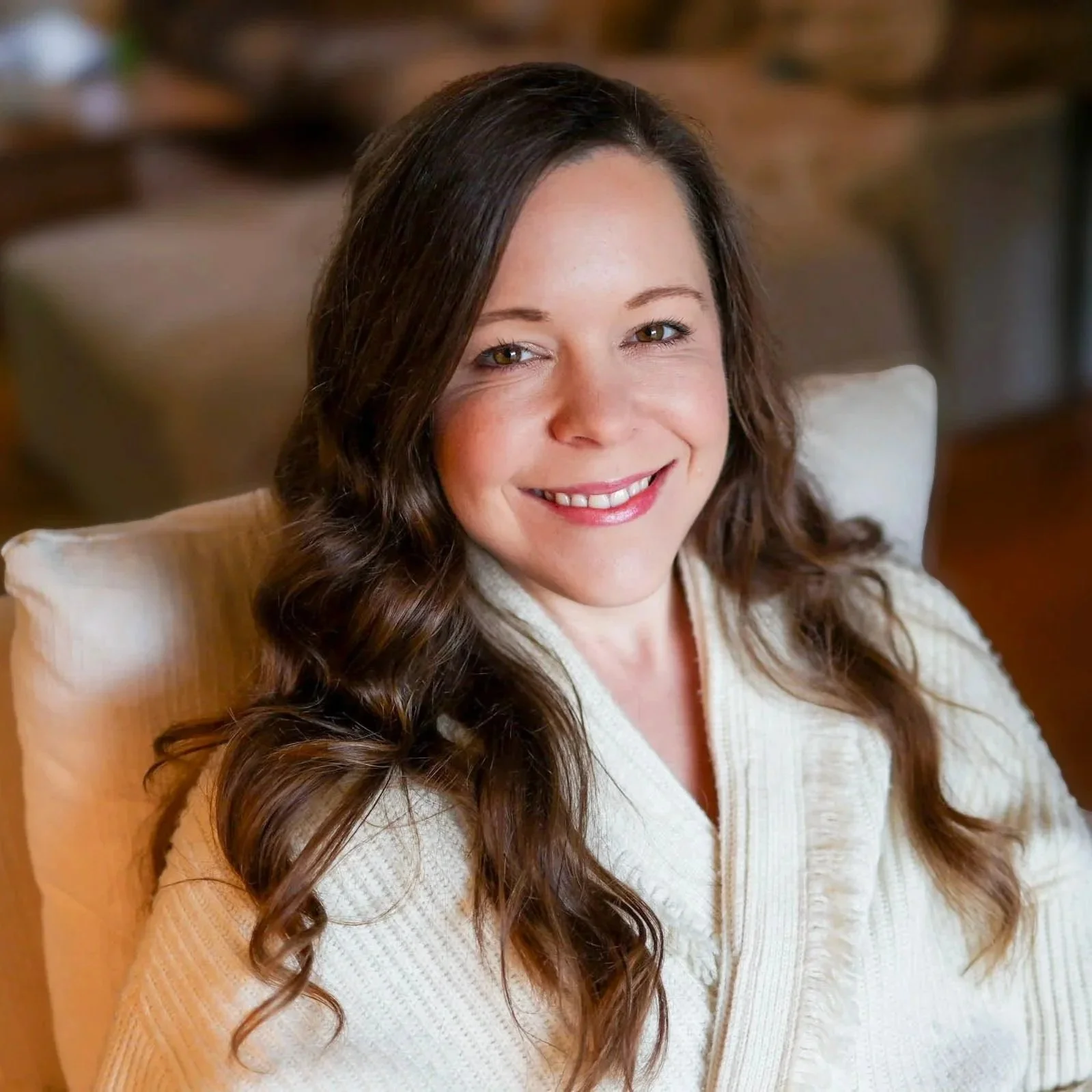 professional photo of Dr. Anna smiling warmly, wearing cozy cardigan, sitting in large chair with blurred background.
