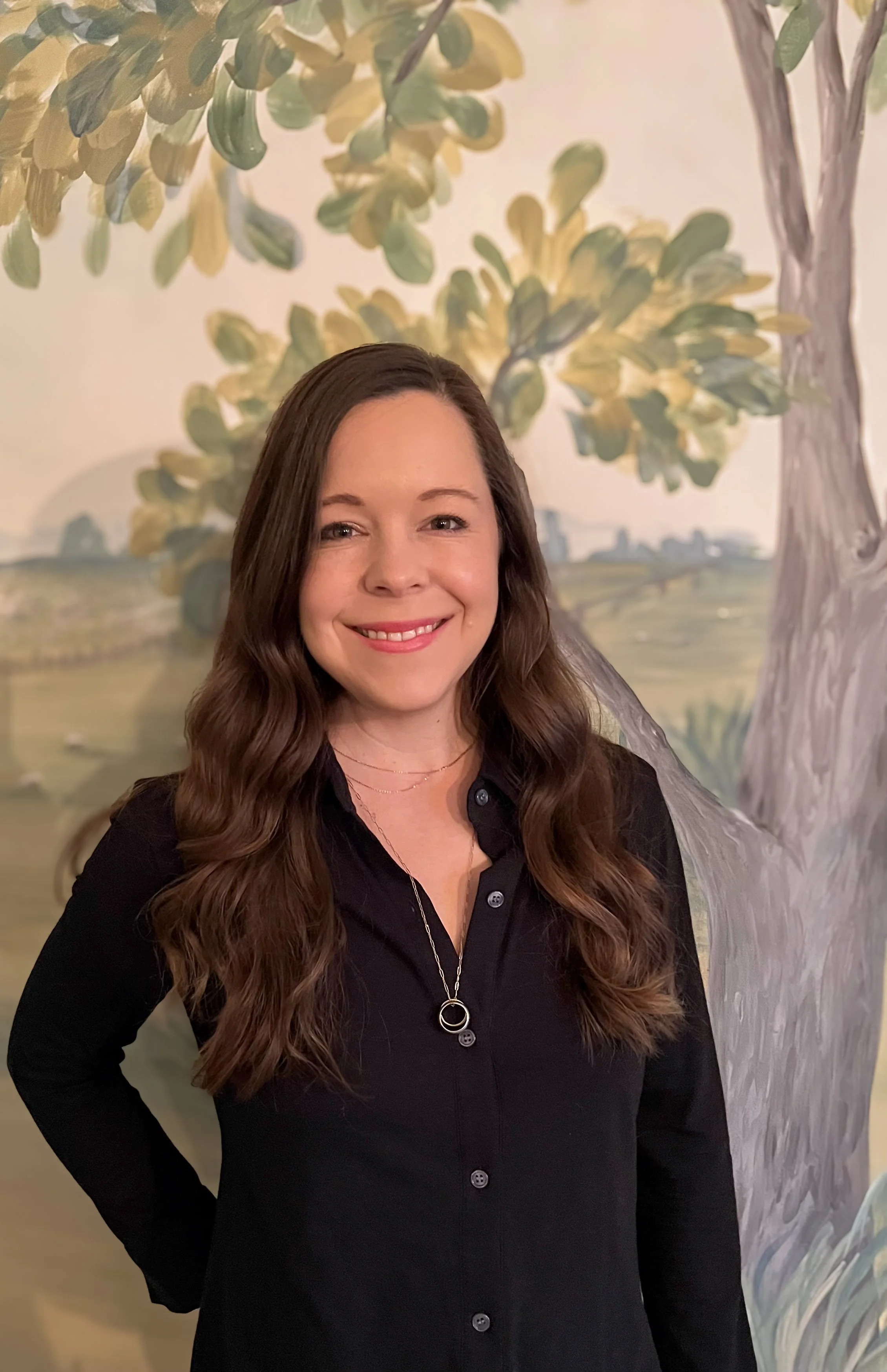 photo of Dr.Anna smiling warmly at the camera, standing in front of mural painting.