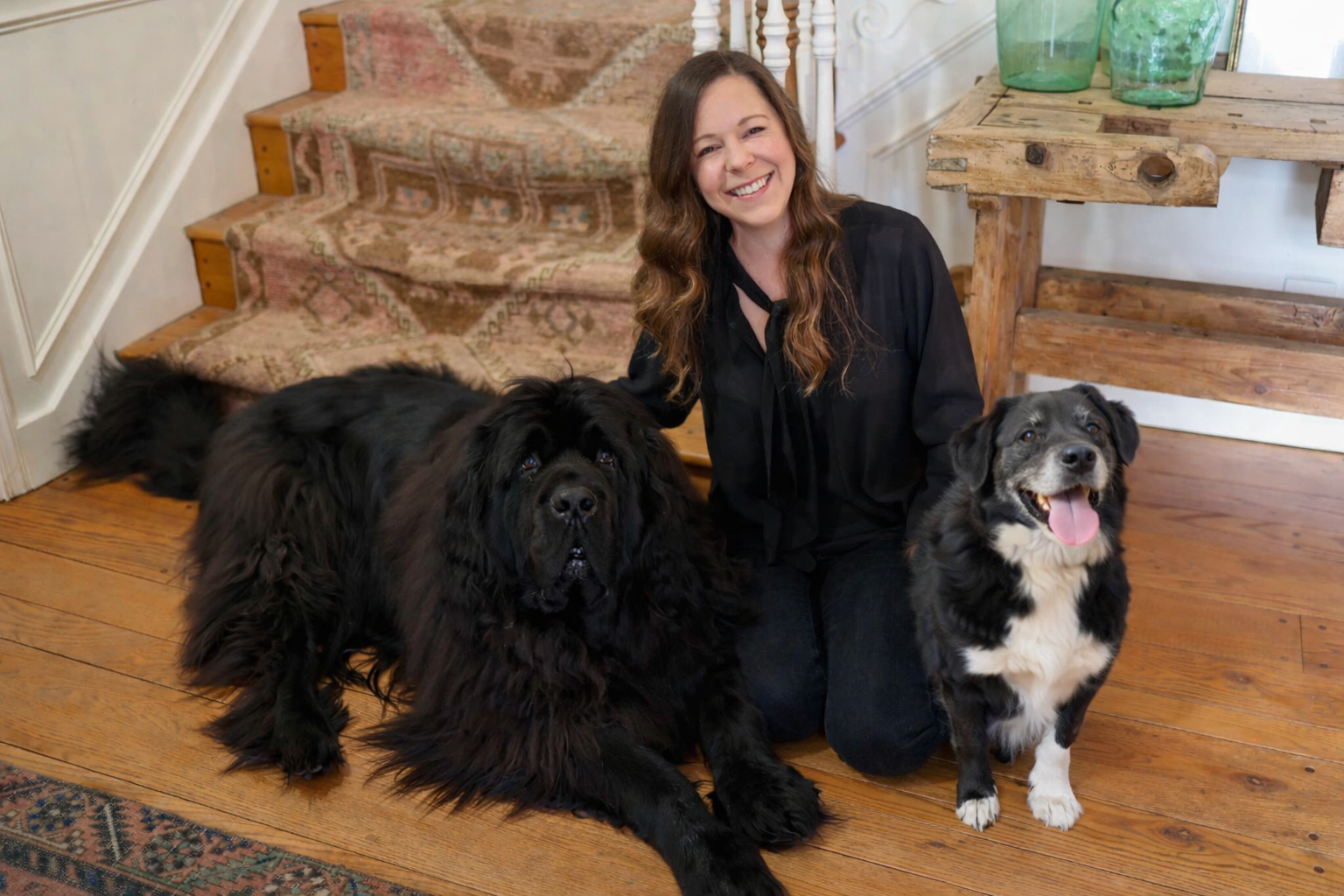 photo of Dr. Anna smiling happily with two dogs at her feet, one very large all black dog, the other smaller black and white dog.