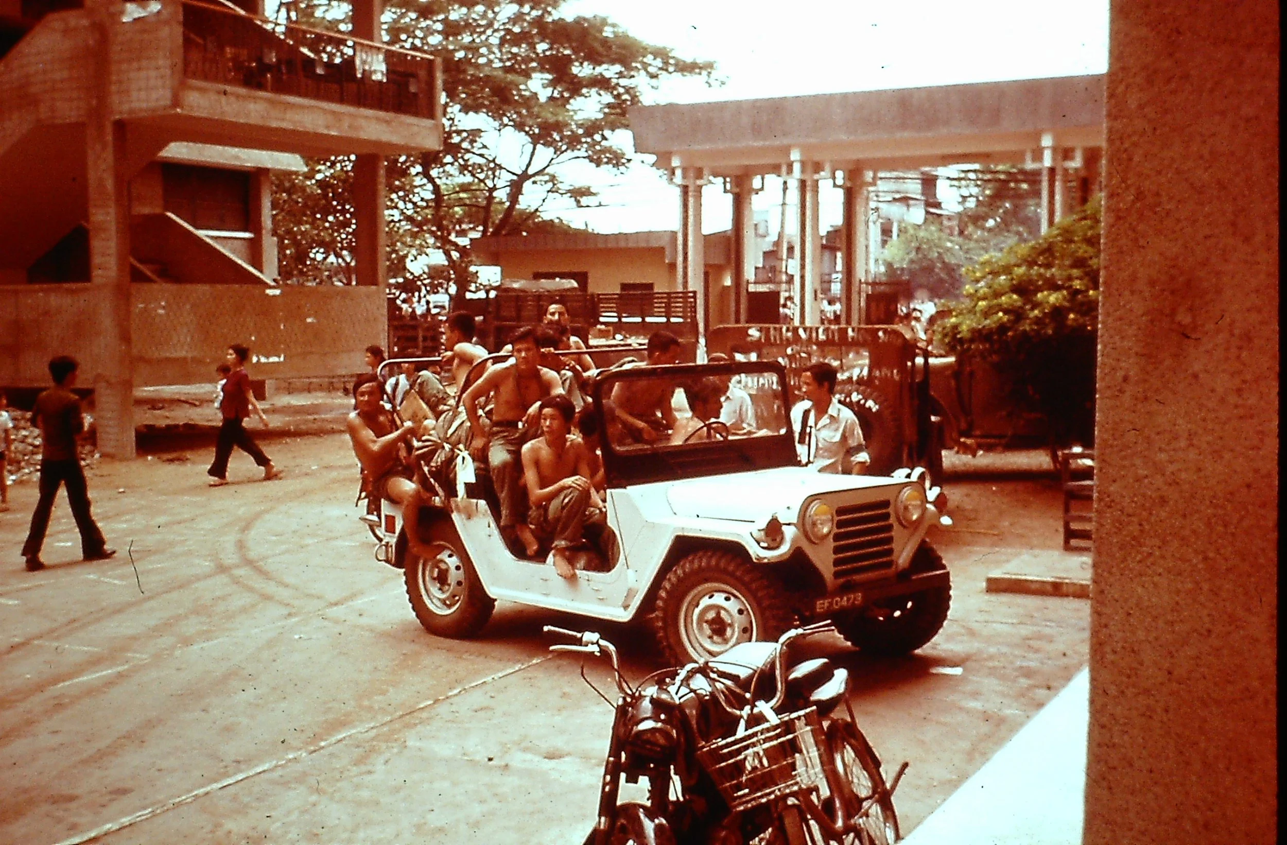 A black and white photo of a small open utility vehicle with several men and women sitting or standing on it, carrying backpacks, on a street in an urban area with buildings, trees, a motorcycle in front, and other pedestrians in the background.