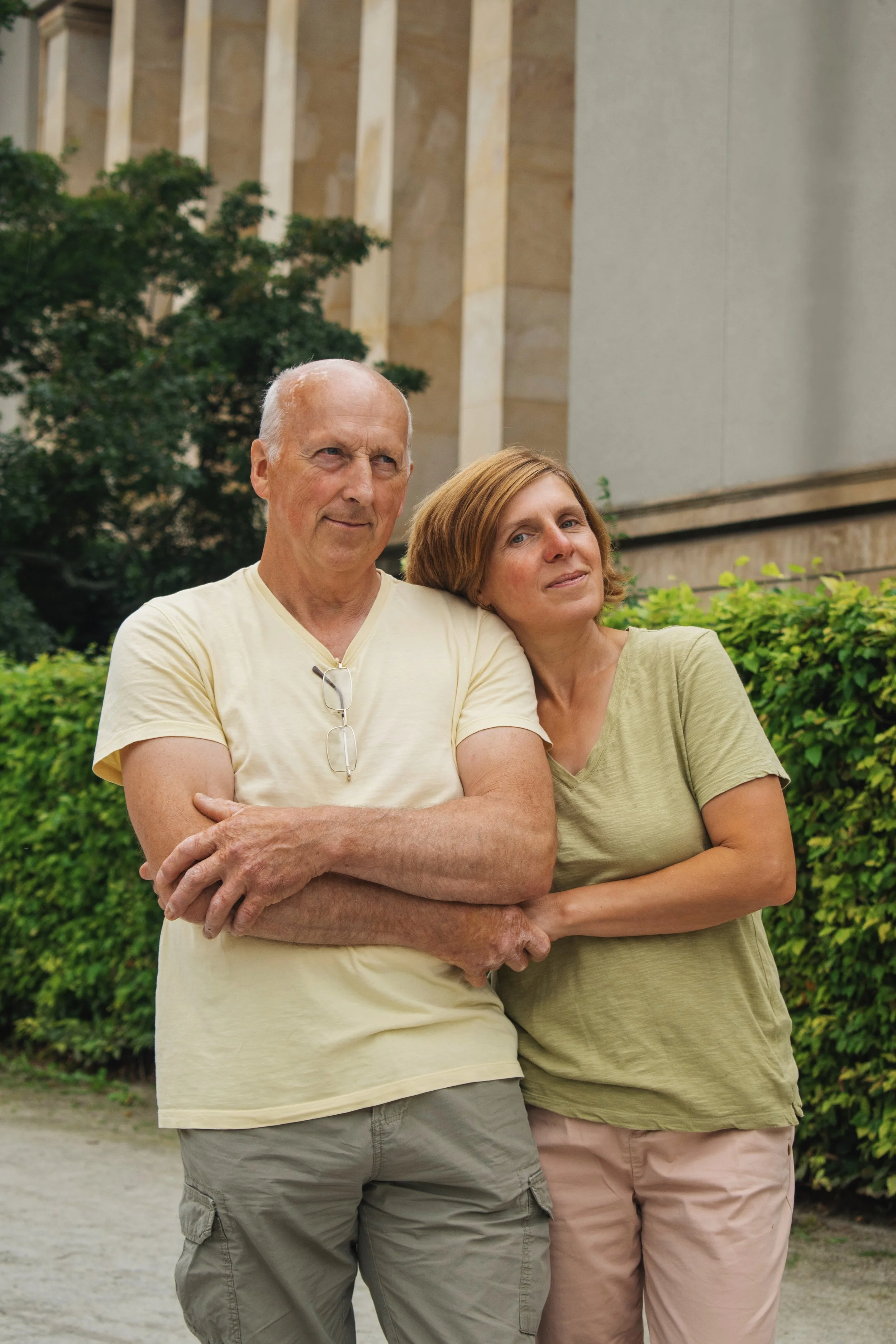 An elderly man and woman stand outside near greenery and a building, with the woman resting her head on the man's shoulder, both looking content.