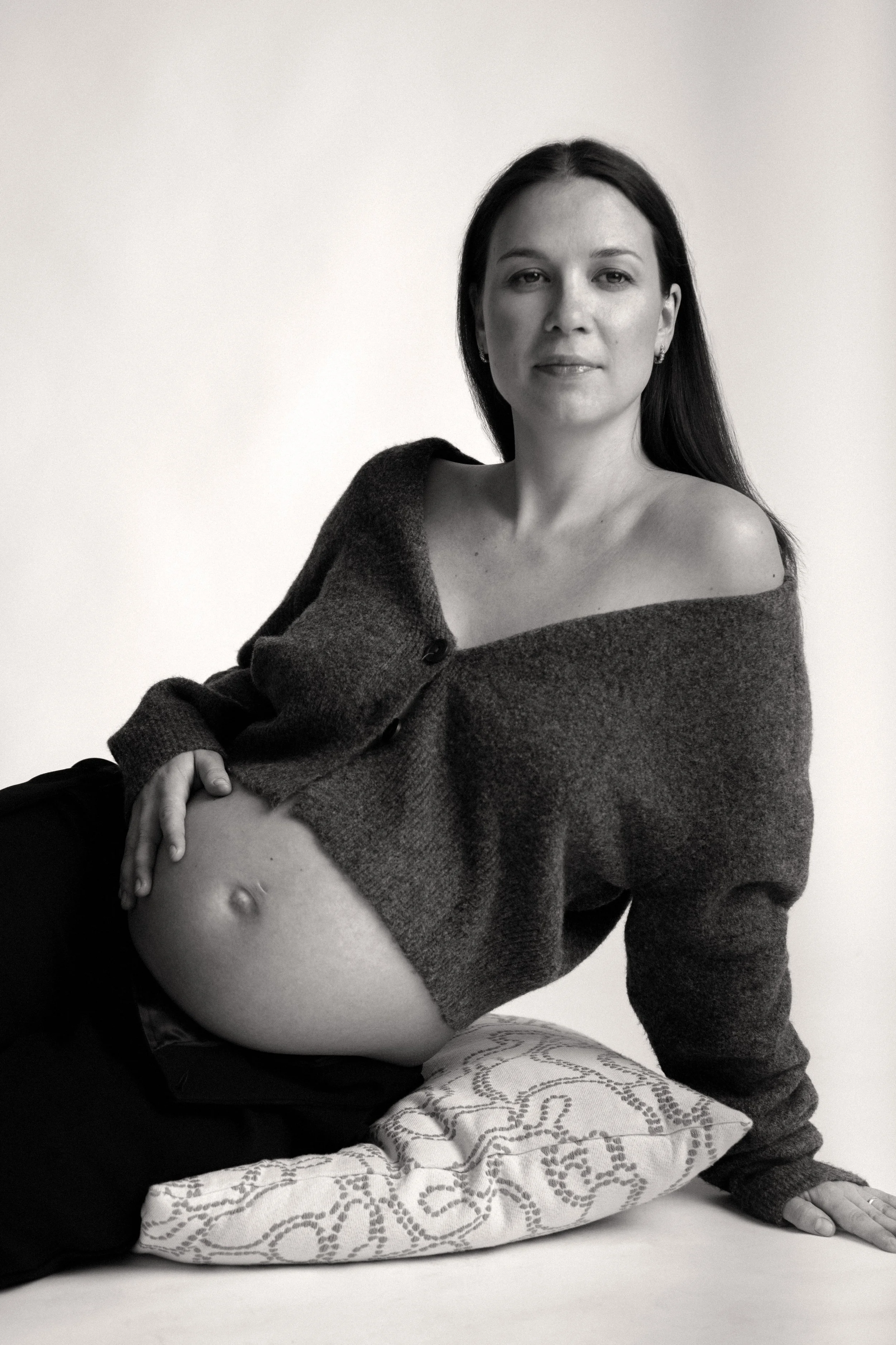 A pregnant woman with long dark hair, wearing a partially open sweater, poses lying on a pillow with her arm stretched out on the ground. She is looking at the camera with a neutral expression.