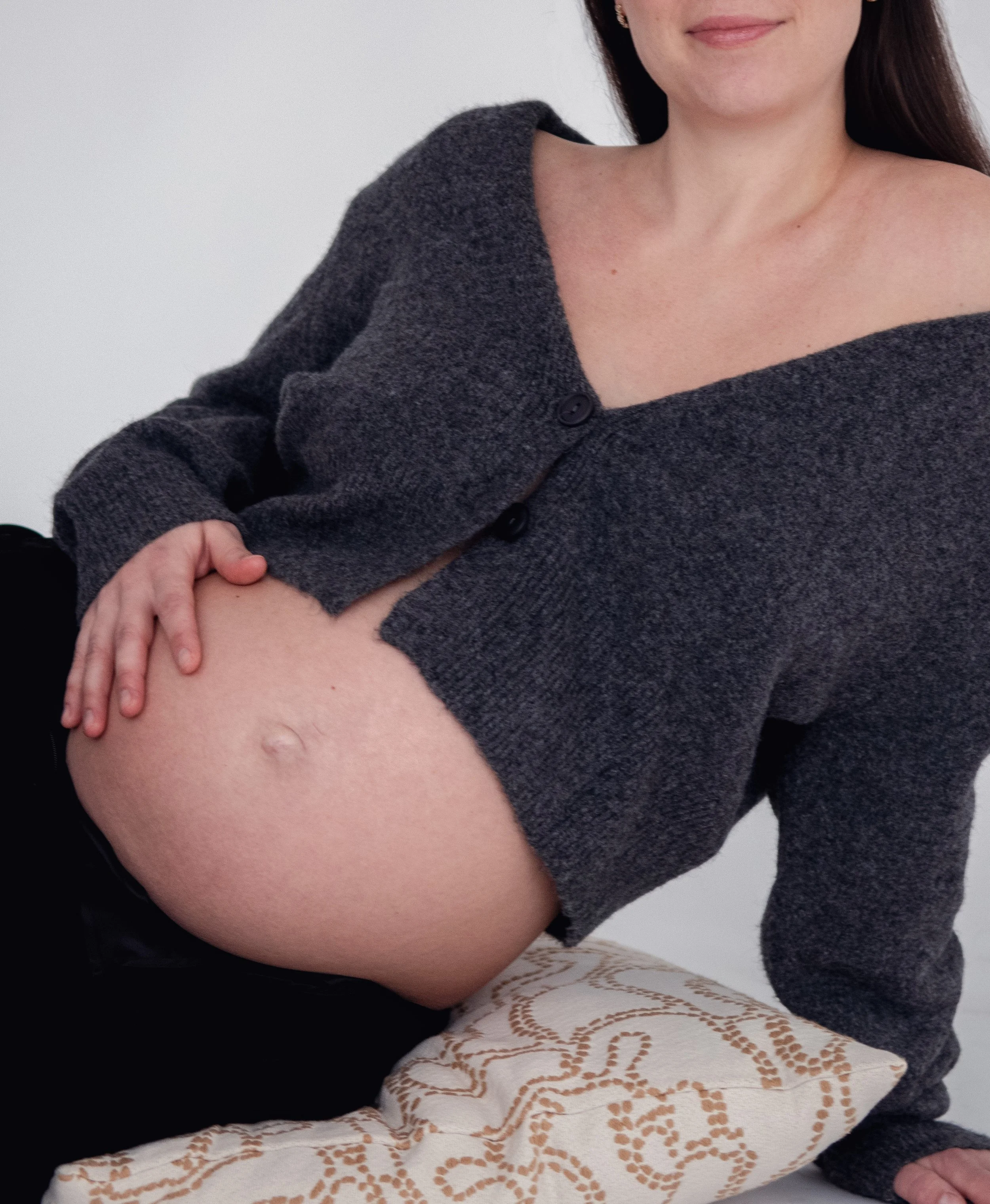 A pregnant woman in a gray cardigan sitting on a white pillow with tan pattern, gently touching her pregnant belly.