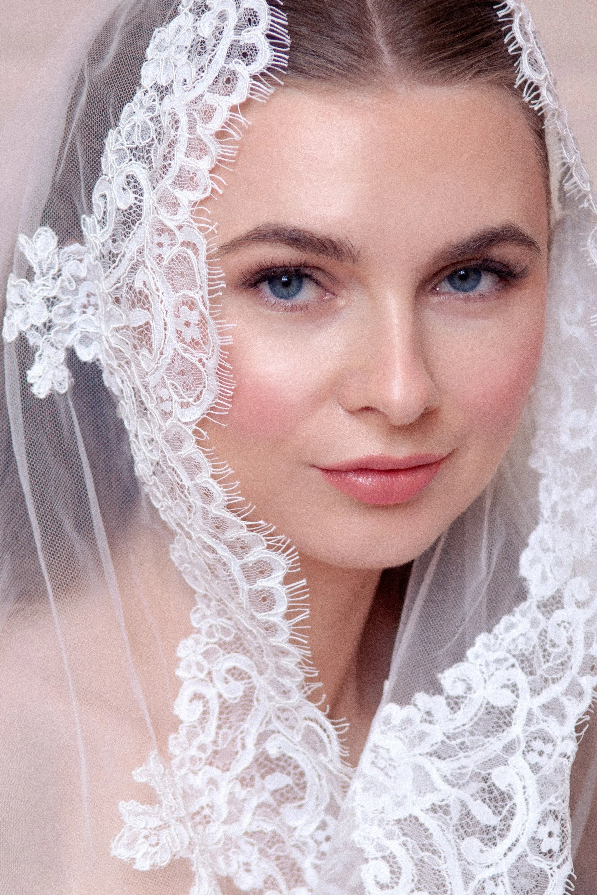 A close up portrait of a bride - woman with blue eyes and light skin wearing a white lace veil with floral embroidery.