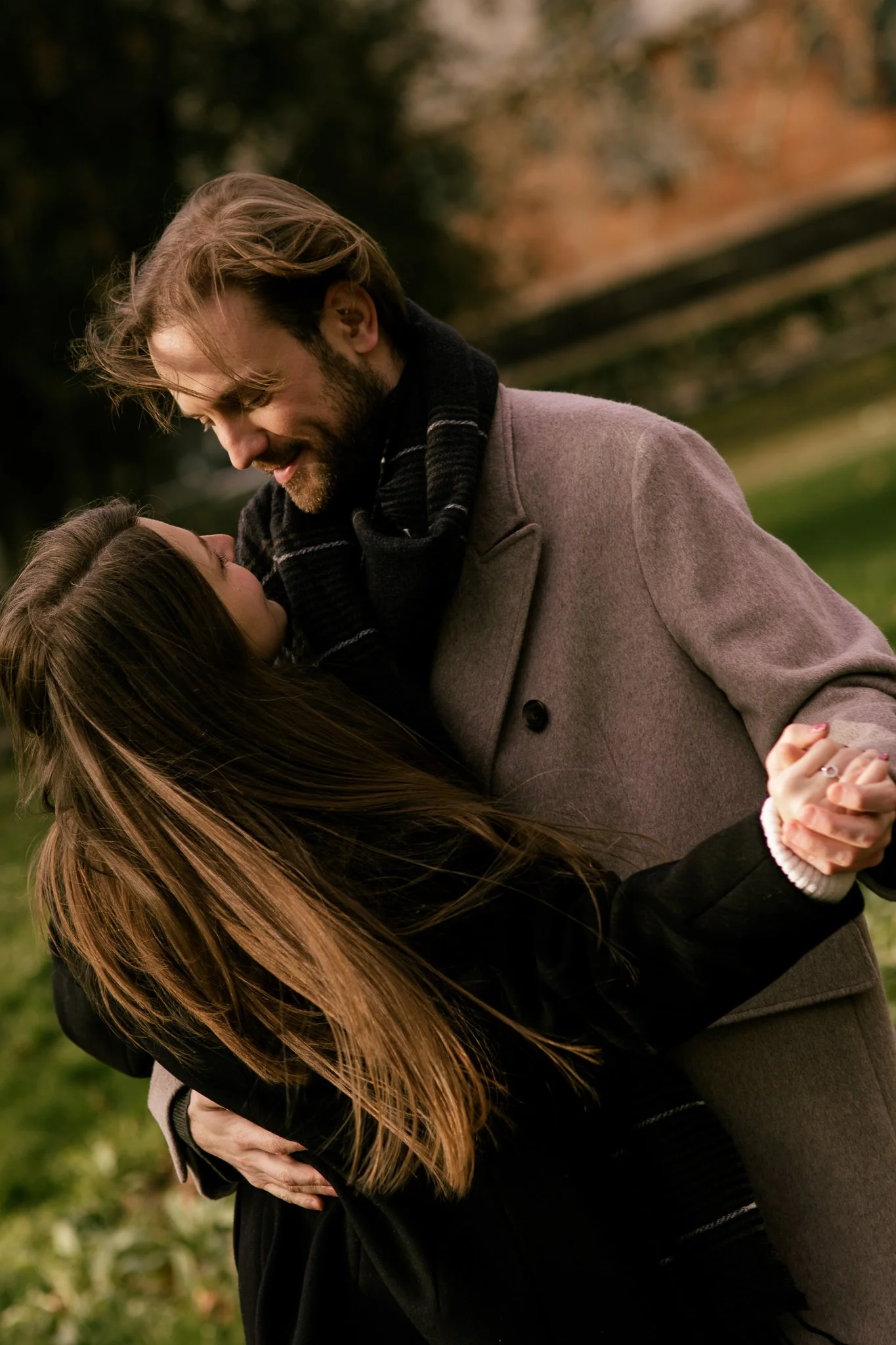 A man and woman dancing outdoors in a park during autumn, holding hands and looking at each other affectionately.