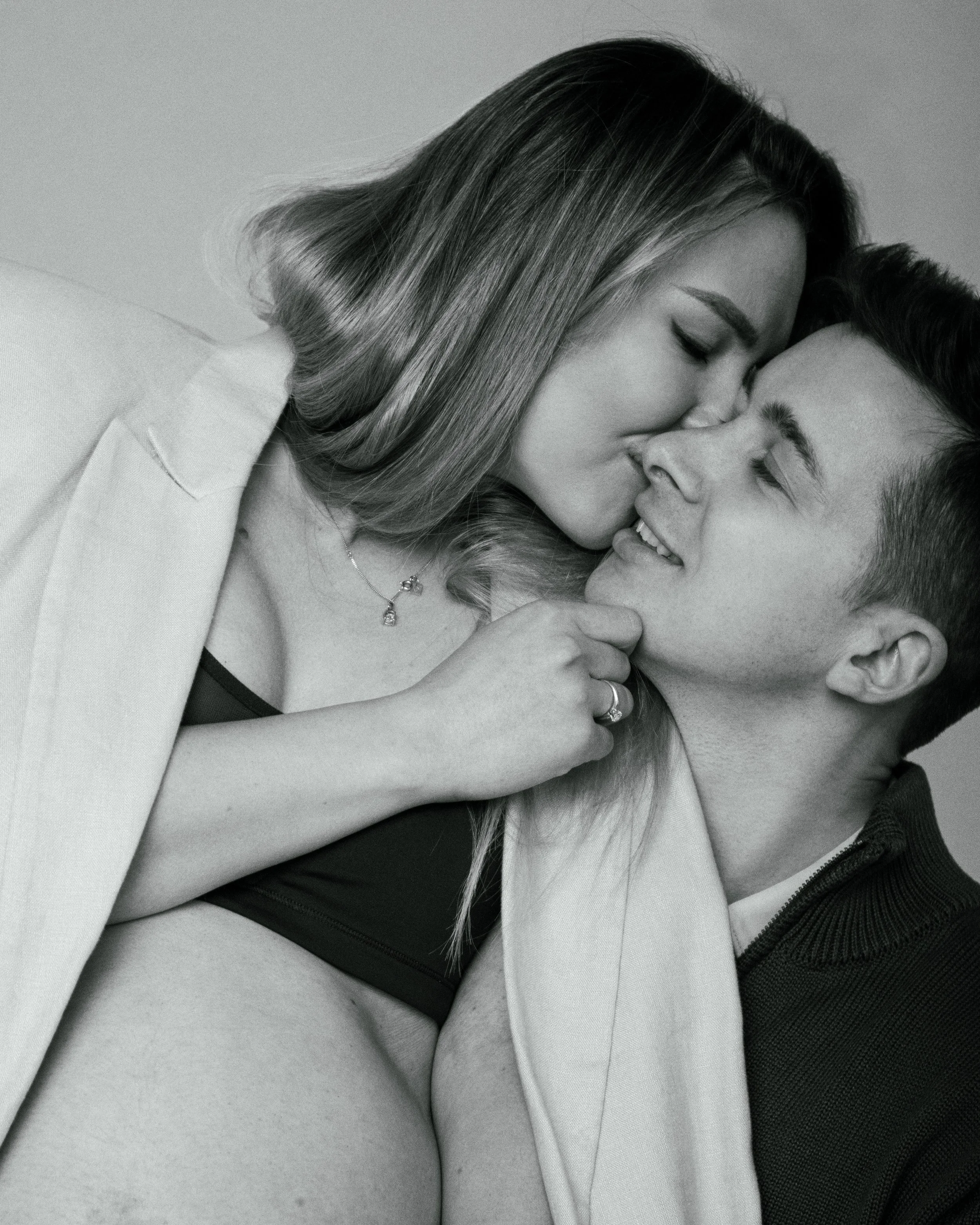 A pregnant woman with long hair kissing a man on the chick, both smiling and looking at each other, in black and white.