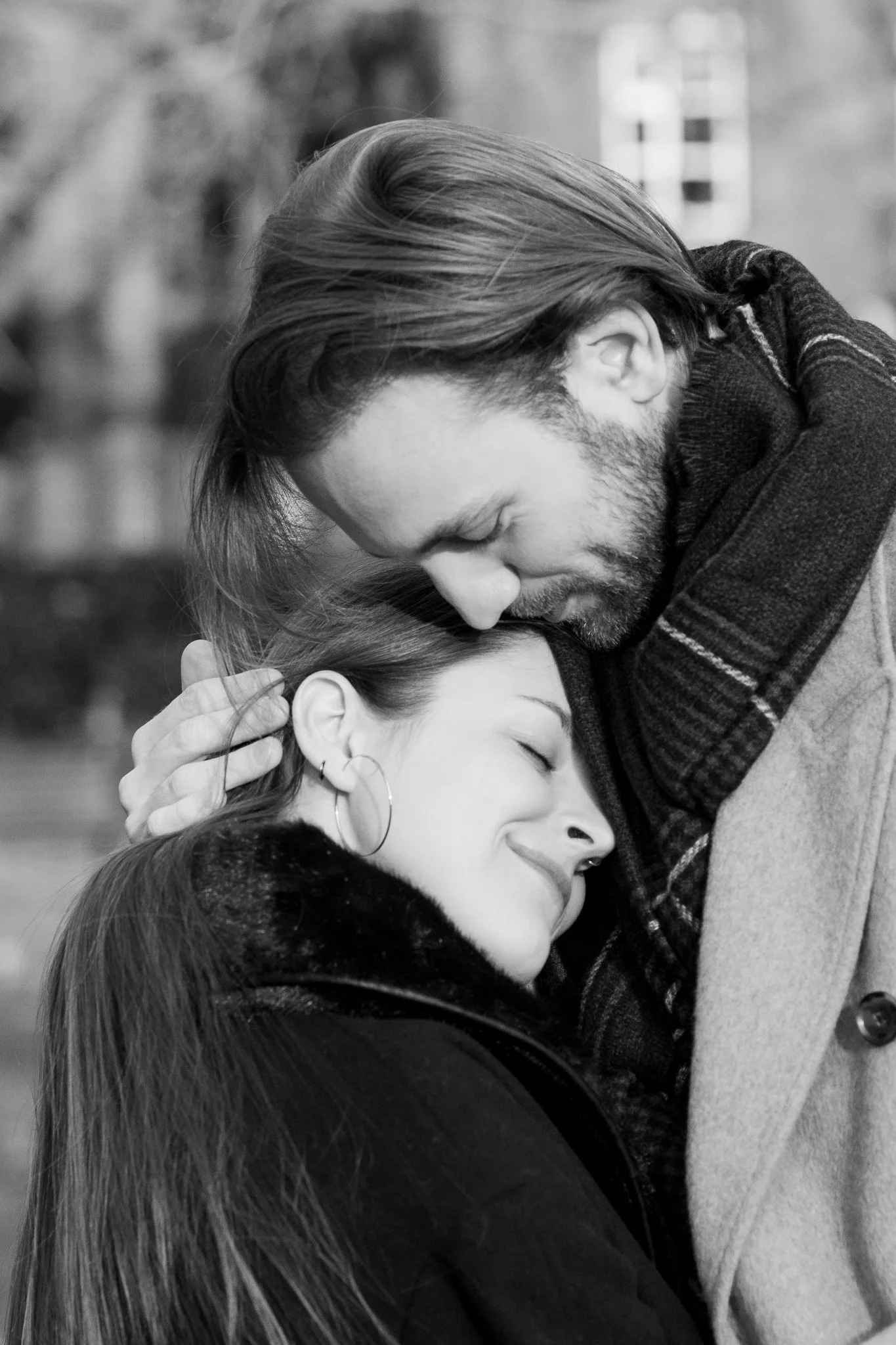 A black and white photograph of a man and woman sharing an emotional hug outdoors. The man has long hair and a beard, and the woman has closed eyes and a gentle smile, wearing hoop earrings.