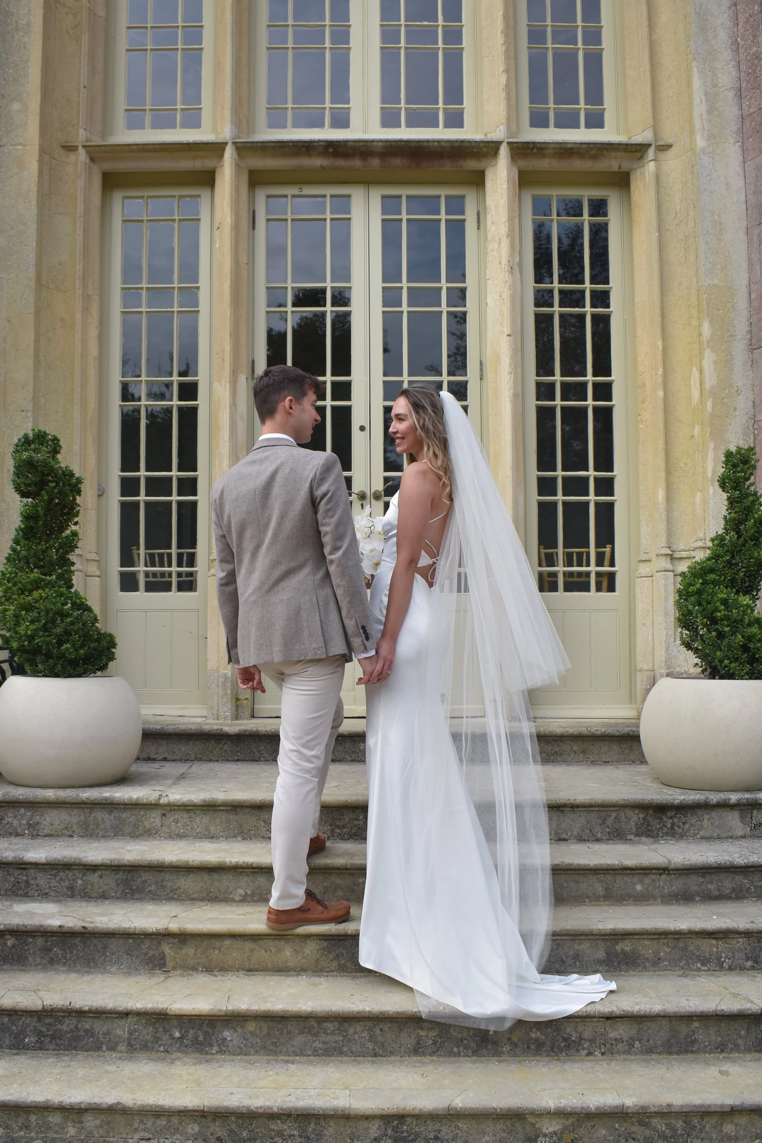 Bride and groom holding hands on the steps outside a historic building with large windows and potted plants.