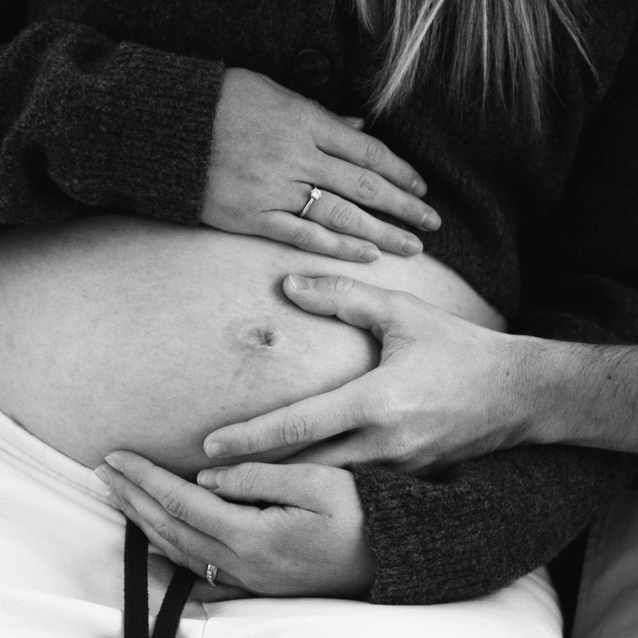 Close-up of a pregnant woman's bare belly being tenderly held by two hands, one on top and one below, in black and white.