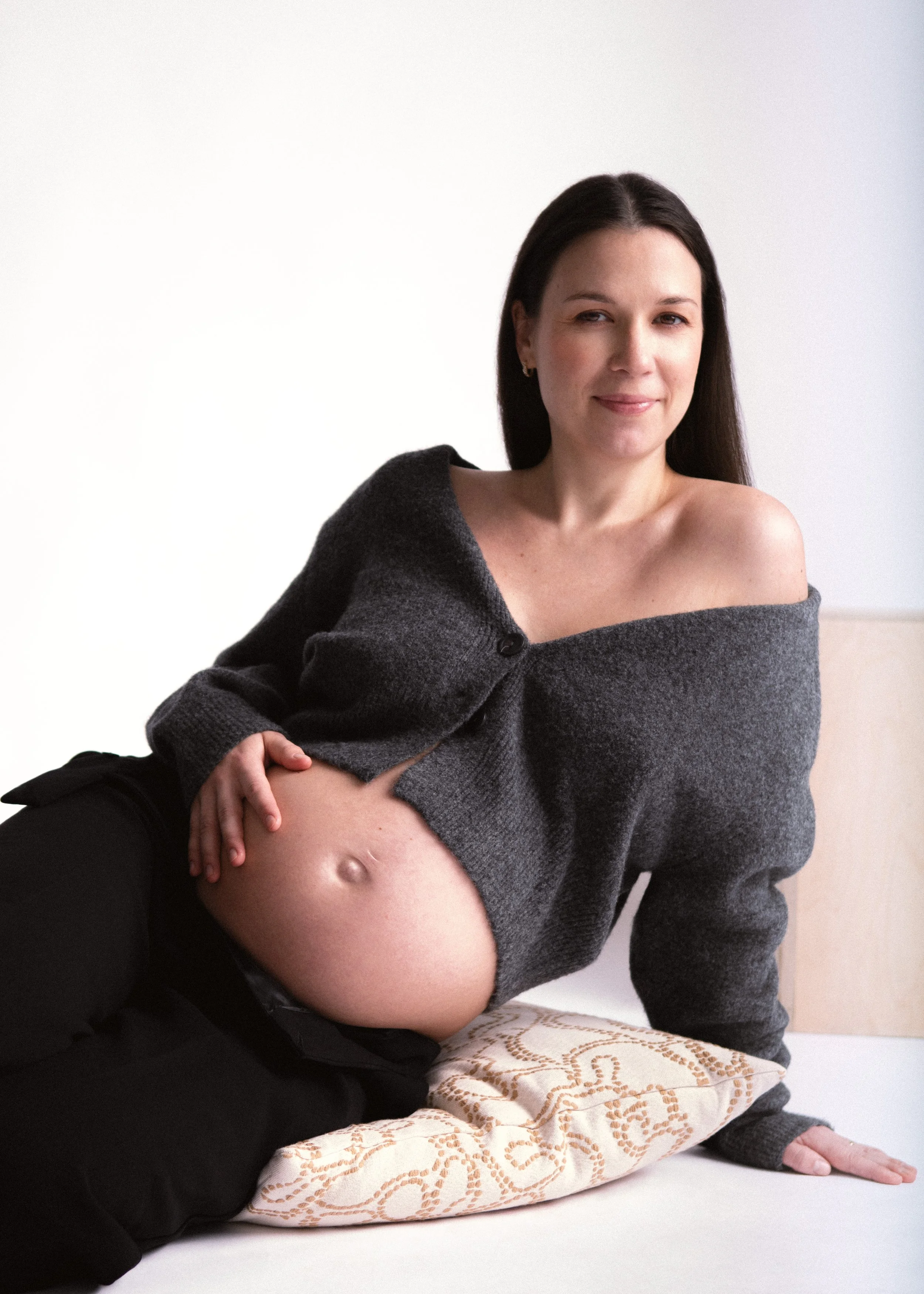 A pregnant woman with dark hair sitting on a bed, wearing a gray off-the-shoulder sweater and black pants, smiling softly at the camera.