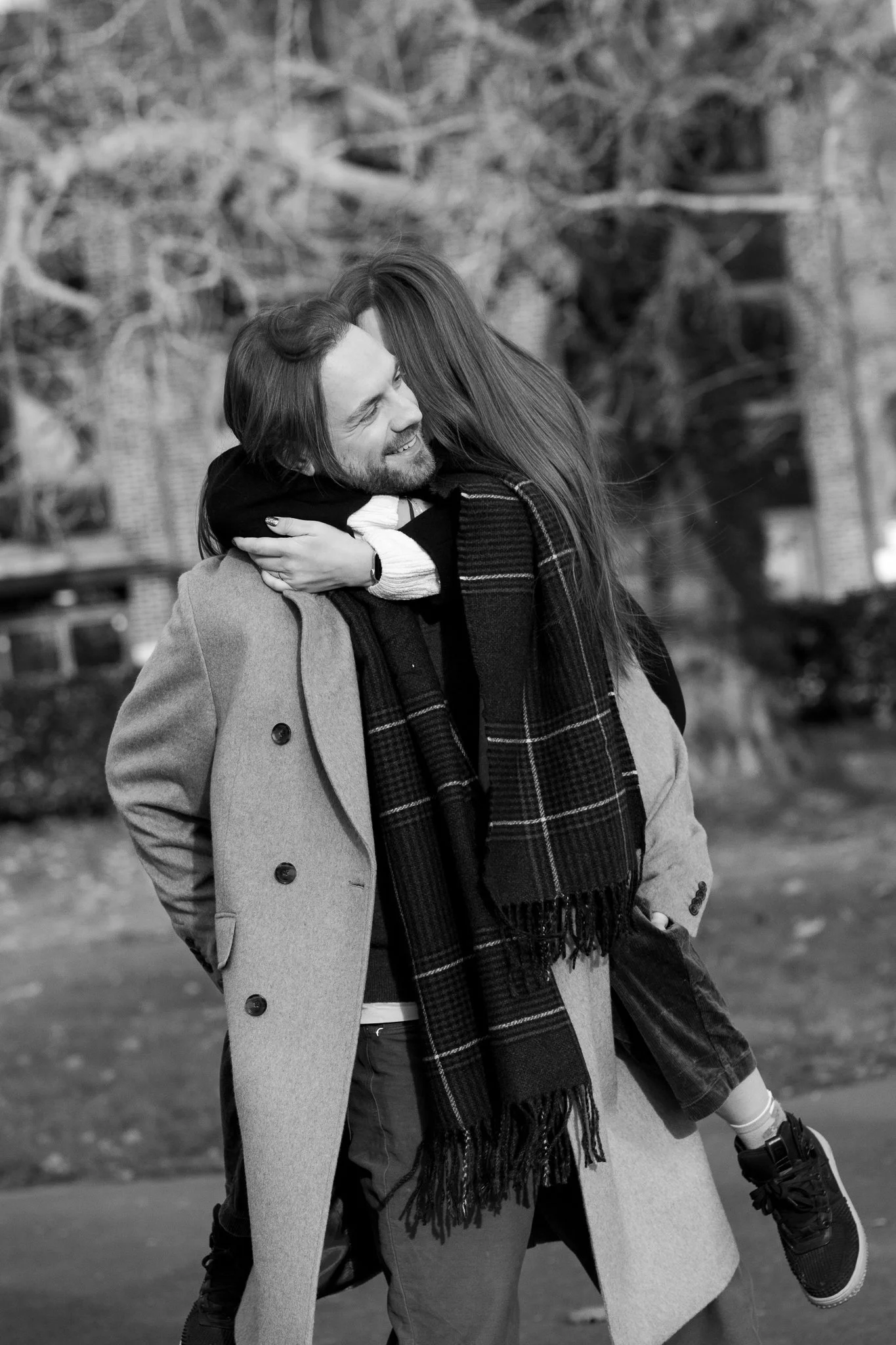 A black-and-white photo of a man giving a piggyback ride to a woman outdoors, both smiling. The man is wearing a coat and the woman is wearing a scarf and sneakers.