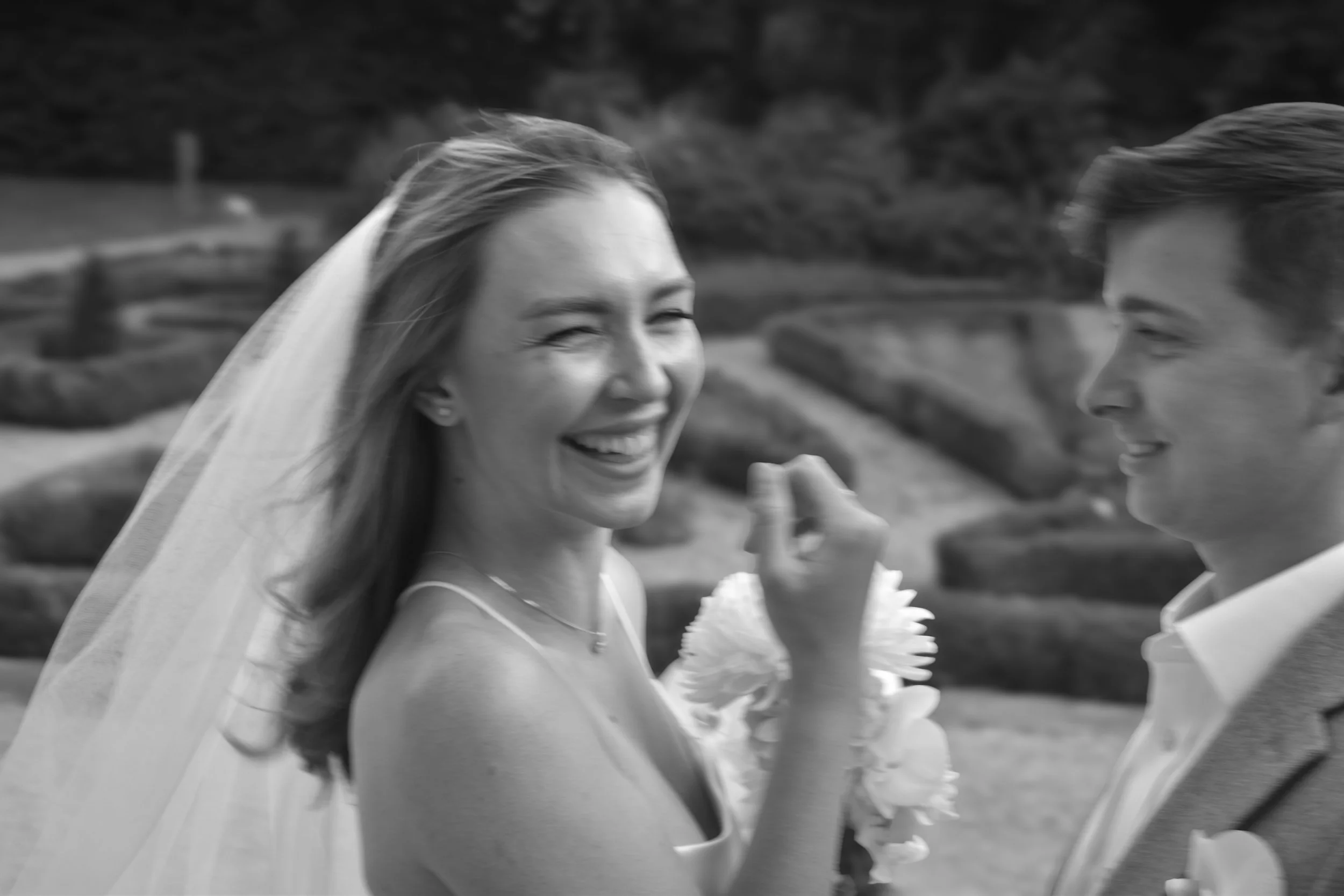 A bride and groom share a joyful moment outdoors, with the bride smiling and holding a bouquet, and the groom facing her, in a black and white photo.