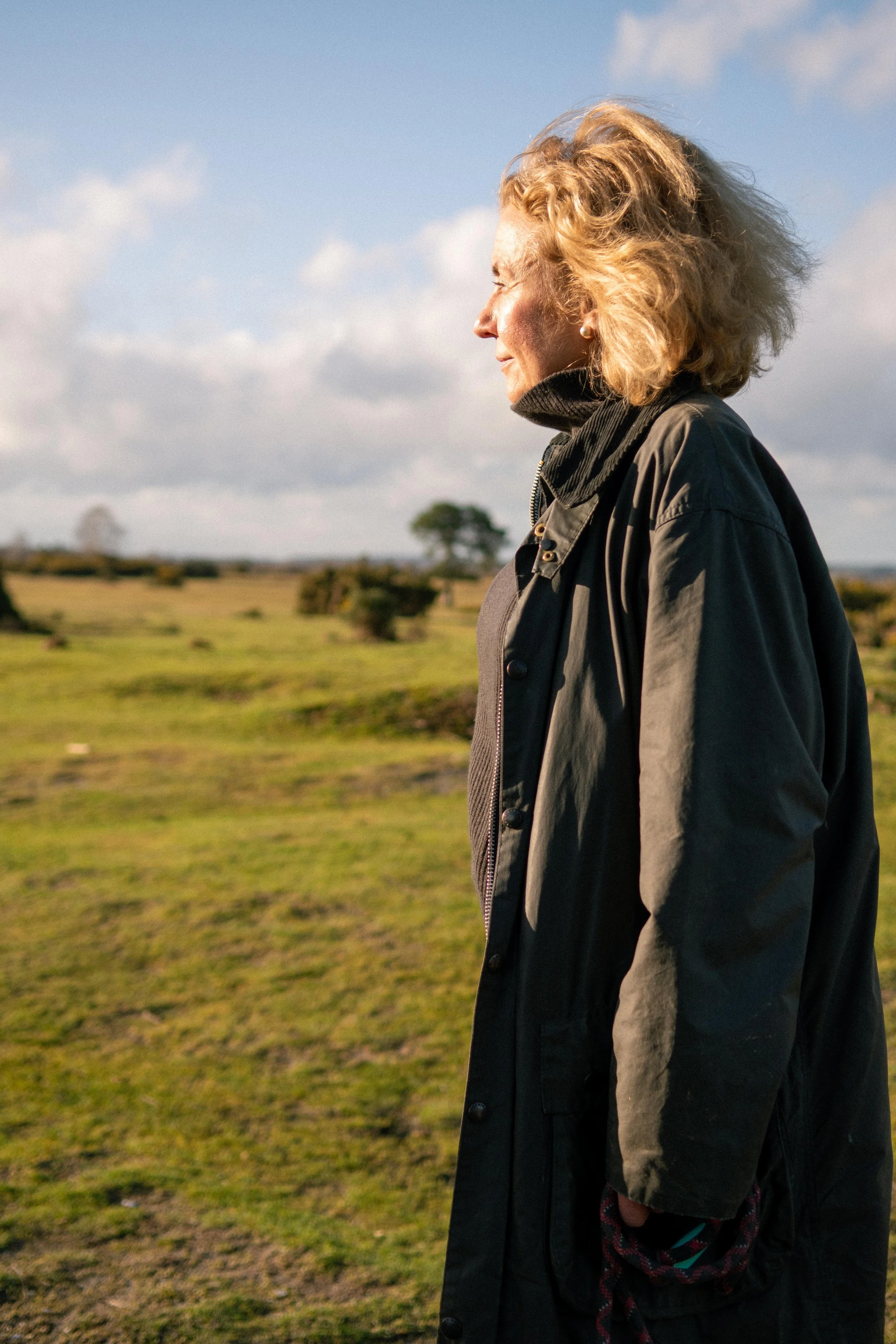 Side profile of a woman standing outdoors in a grassy field with some trees and a cloudy sky in the background. She has short blonde curly hair and is wearing a dark jacket.
