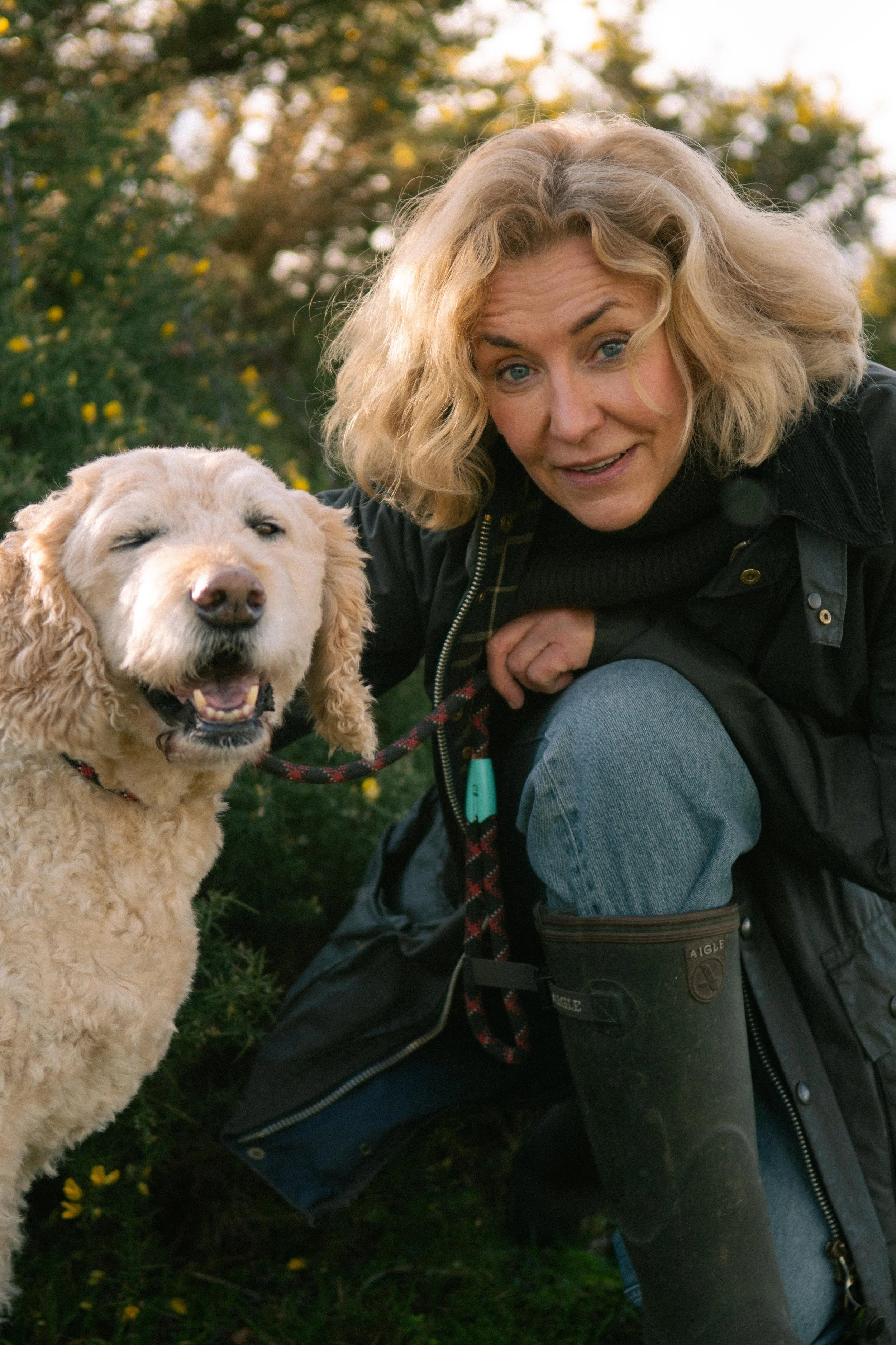 A woman with shoulder-length blonde hair crouching outdoors next to a happy golden retriever dog, holding its leash, with greenery and trees in the background during fall.