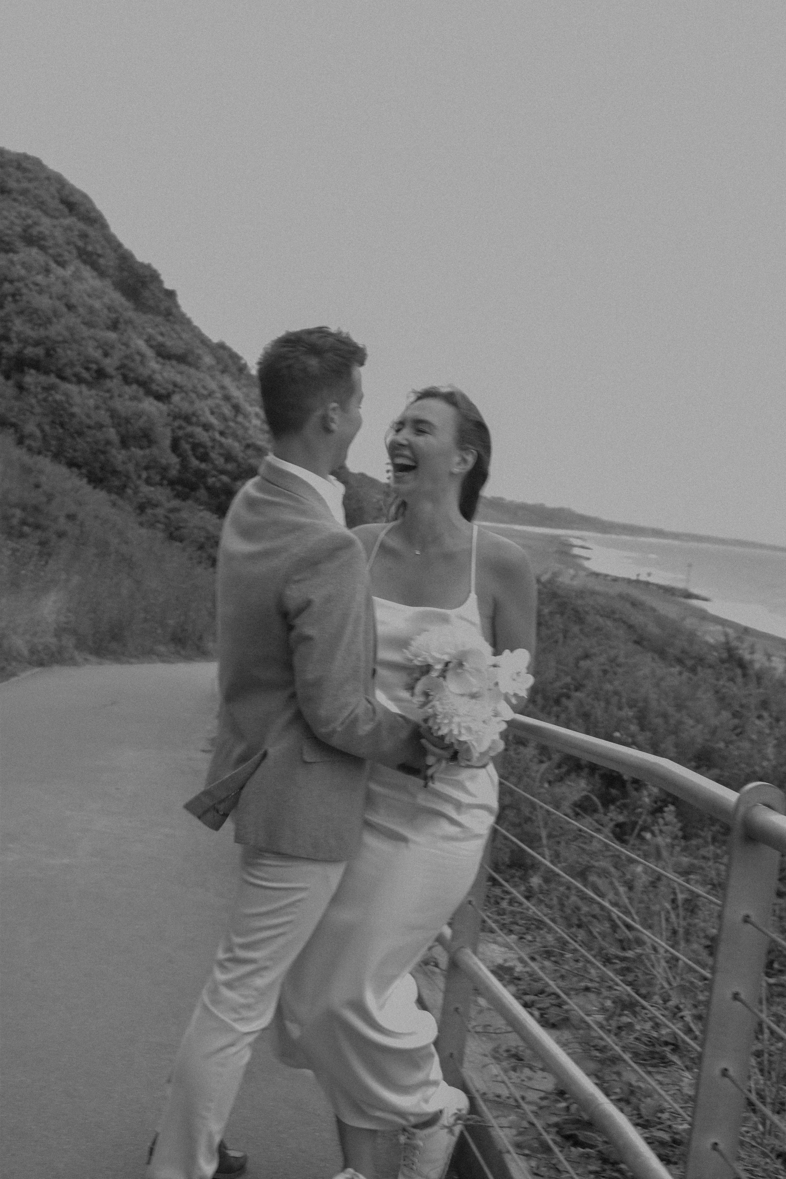 A couple in wedding attire is smiling and enjoying a moment together outdoors on a coastline, with a scenic view of water and greenery in the background.
