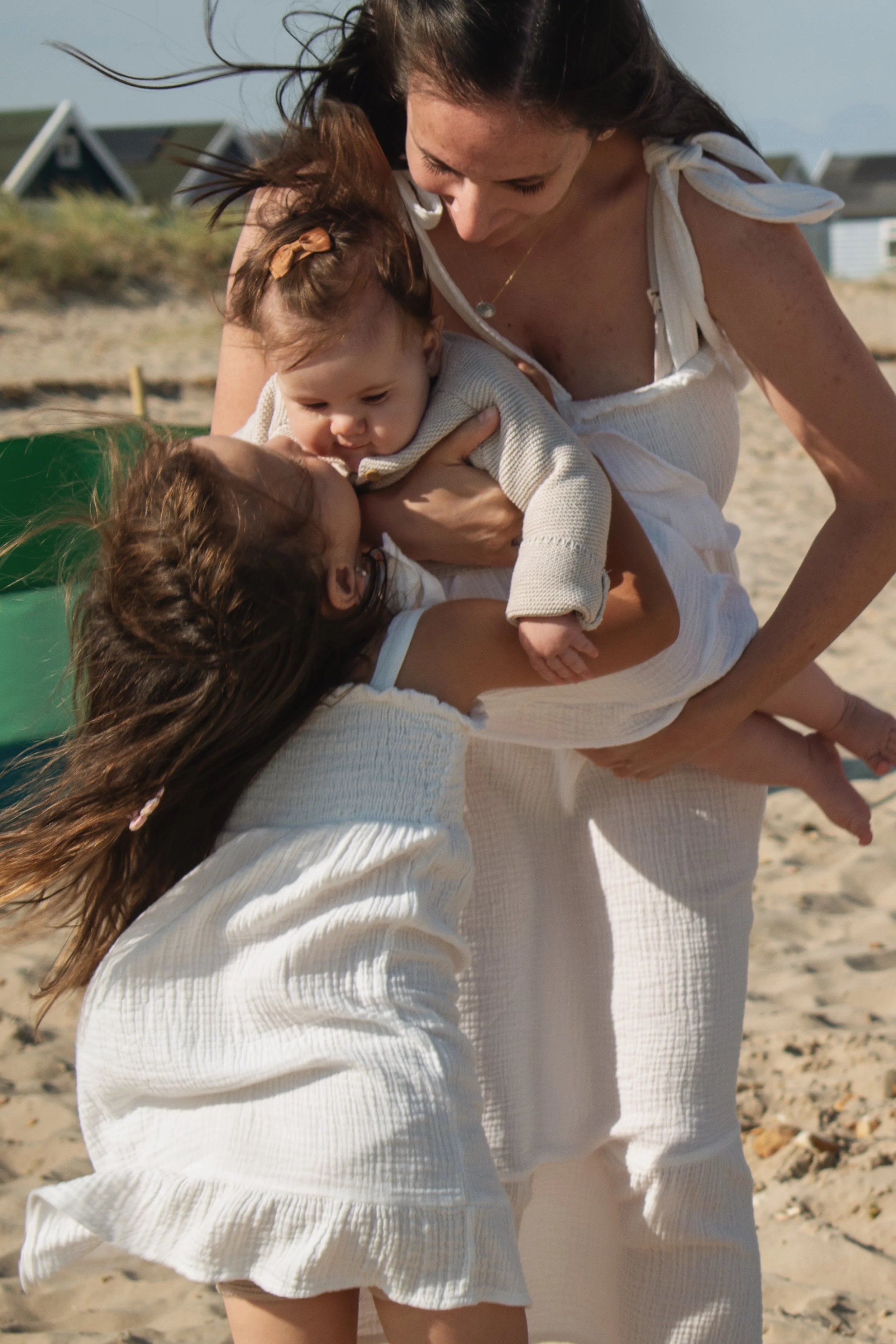 A woman is lifting two young girls on a sandy beach, smiling and playing with them, children are dressed in white, and background shows houses and blue sky.
