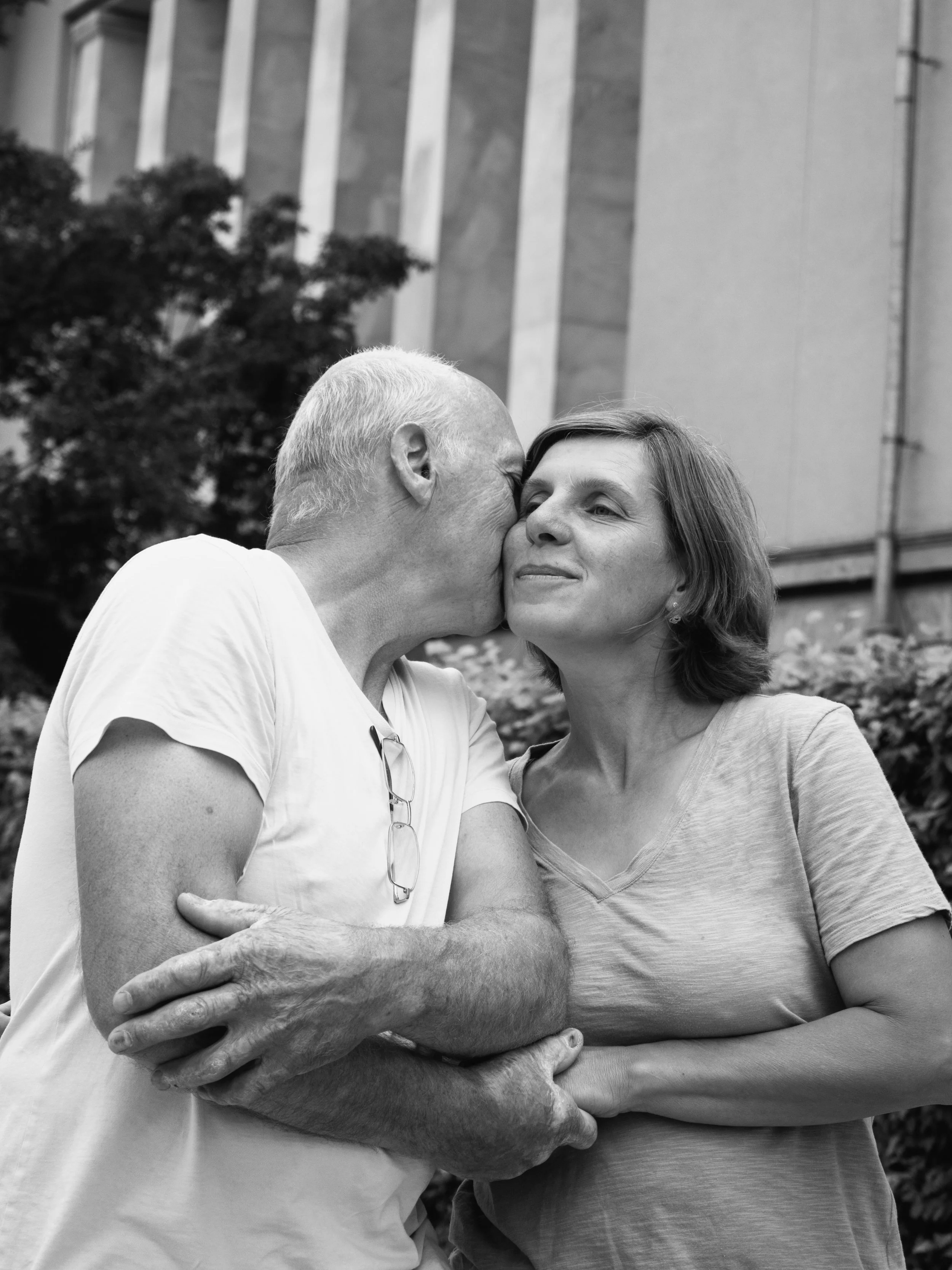 An elderly man kisses a woman on the cheek in front of a building, with greenery in the background.