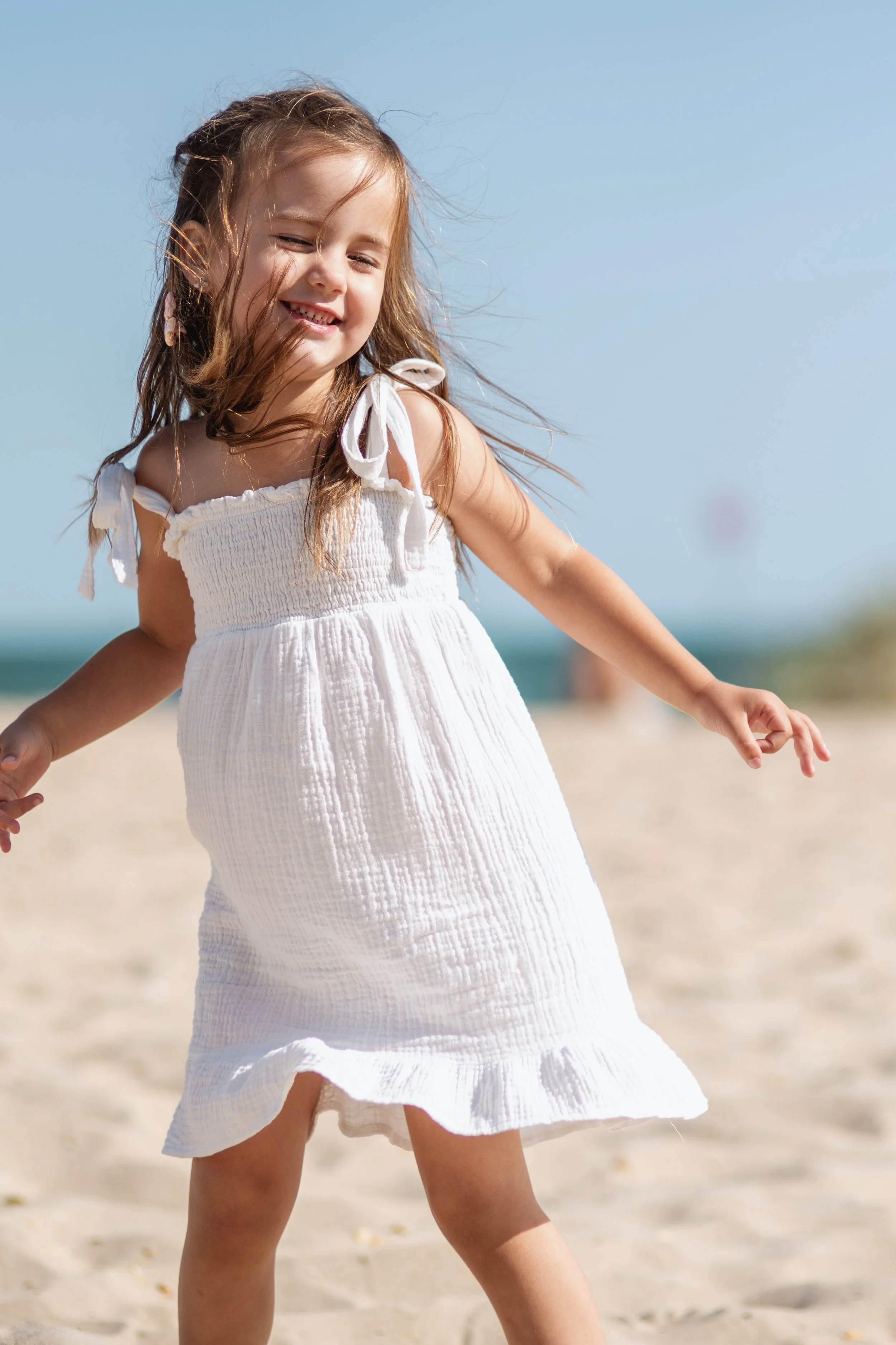 A young girl in a white sundress playing on a sandy beach with the ocean in the background.