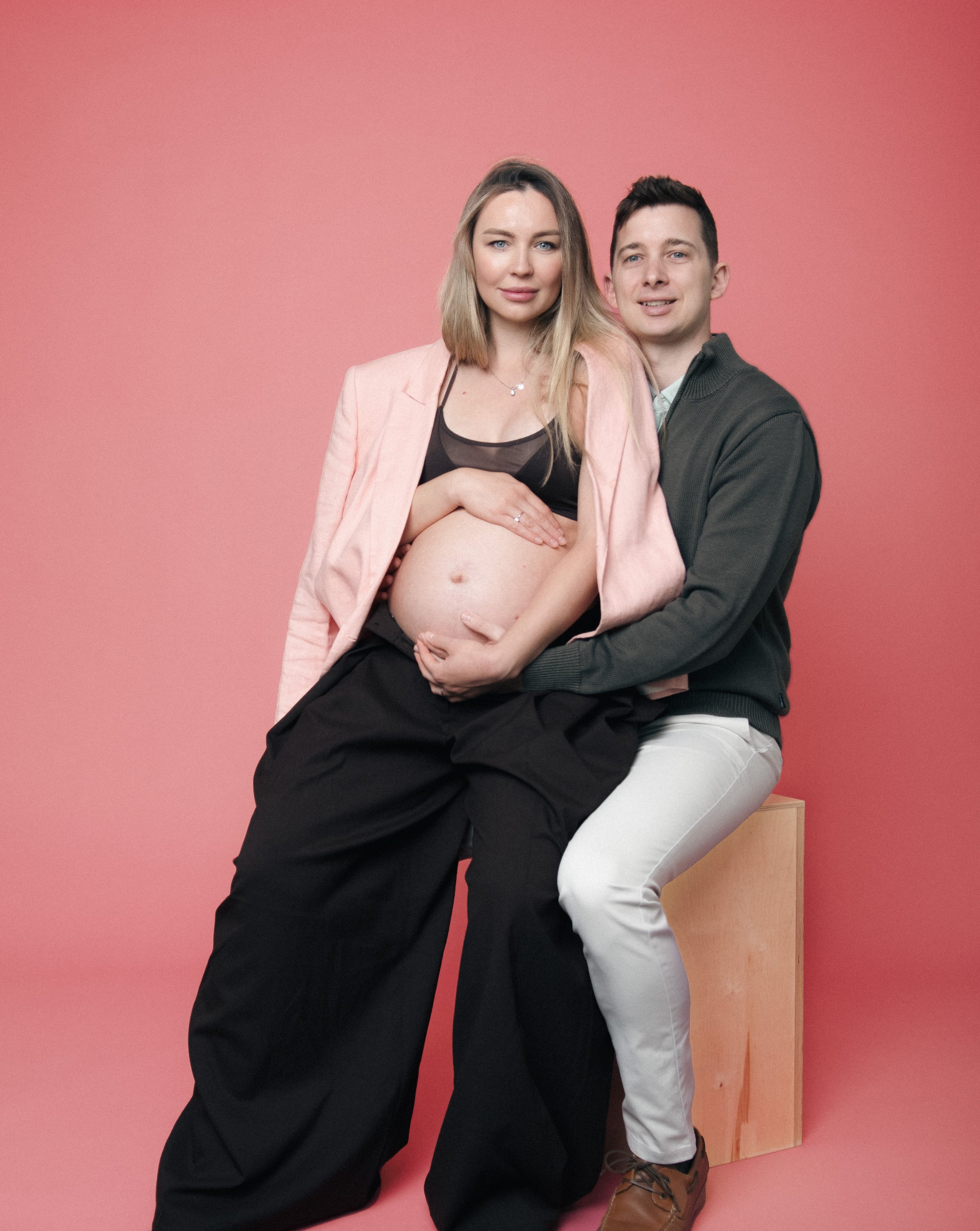 A pregnant woman in brown trousers and a pink blazer, sitting on a husband's lap, who is sitting on a wooden block. The man in a dark sweater and white pants kneeling and holding her belly, against a pink background.