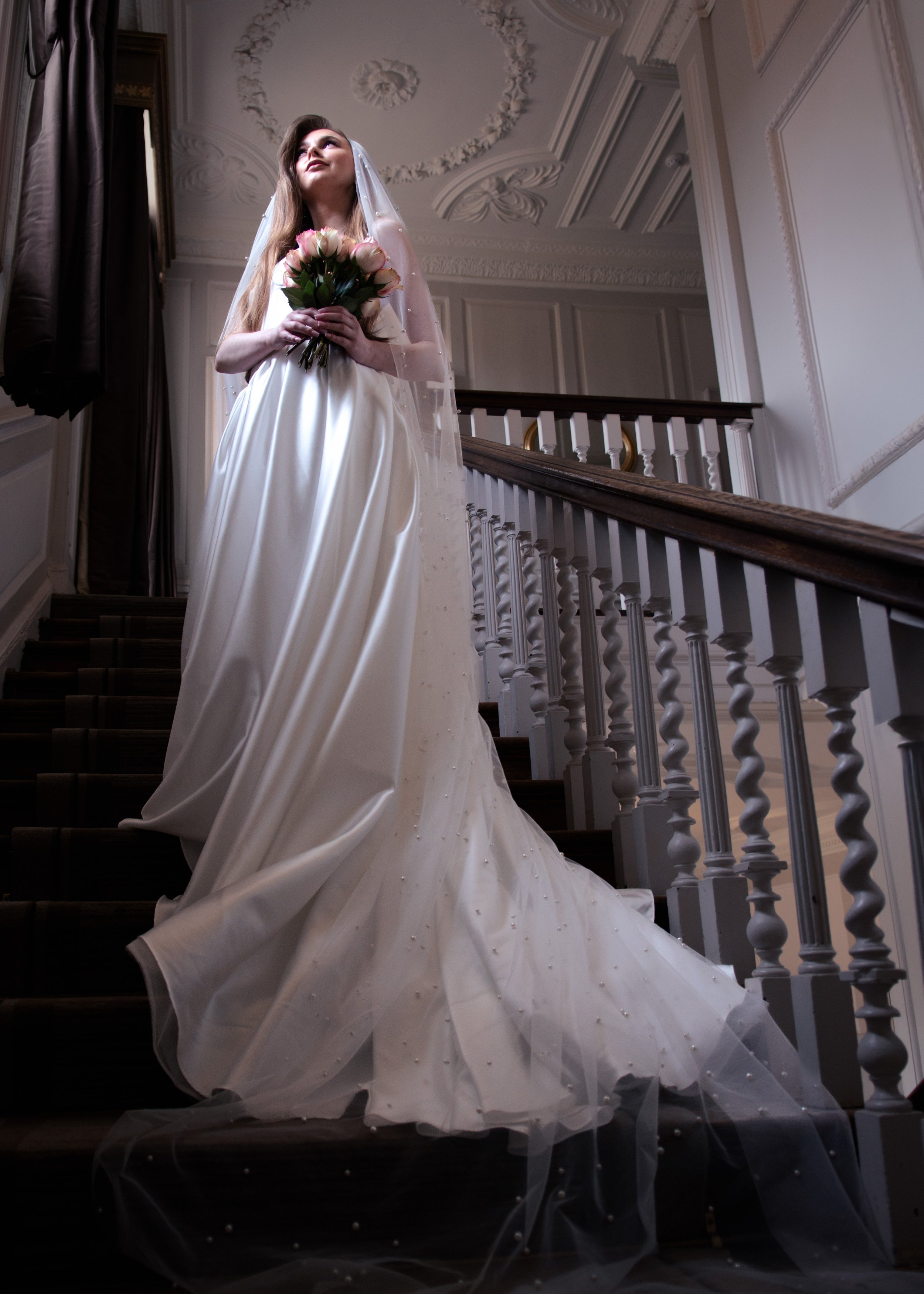 A bride in a long white wedding gown with a veil, holding a bouquet of pink roses, standing on a staircase inside a decorated elegant building.