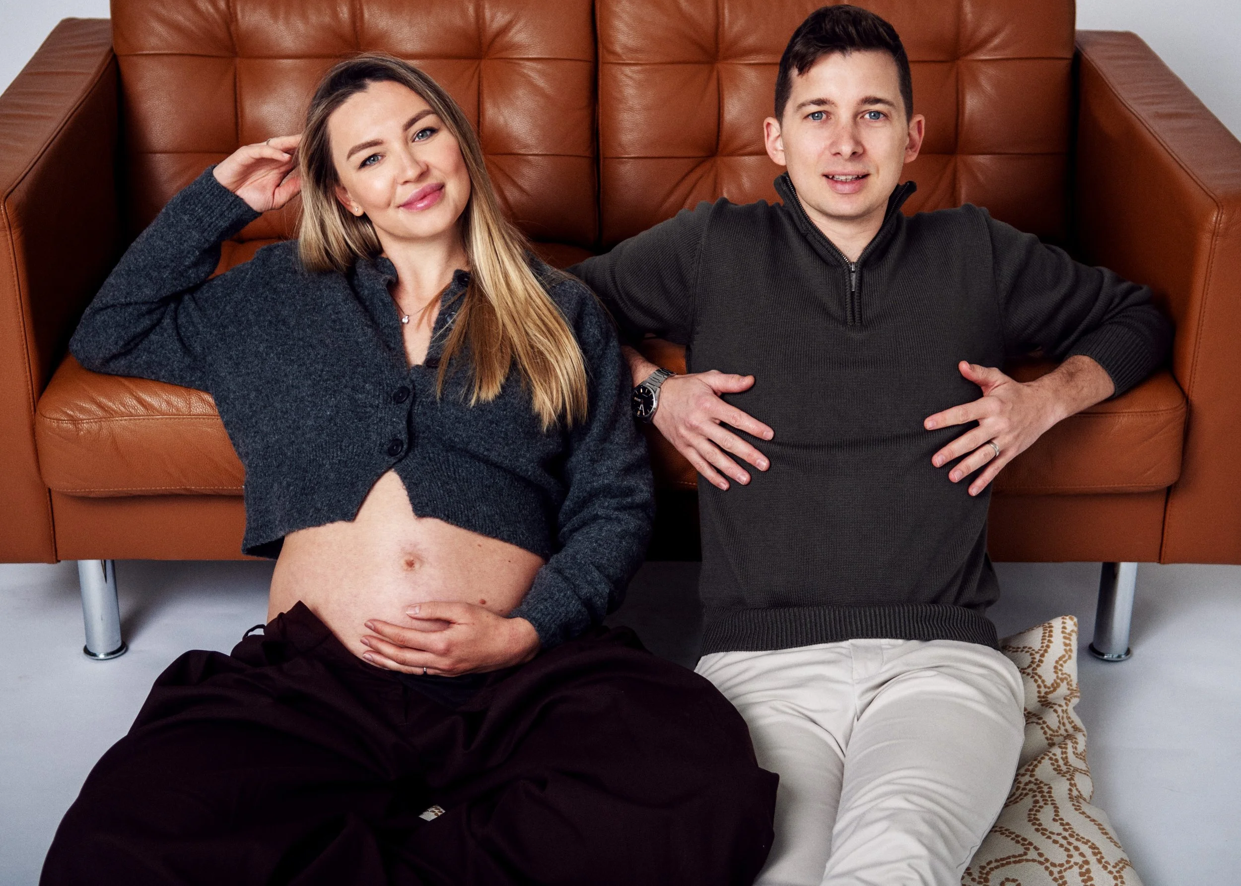 A pregnant woman and a man sitting on the floor, leaning against a brown leather sofa, in a casual indoor setting. The woman is touching her belly with one hand and smiling, while the man has his hands on his stomach and is looking at the camera.