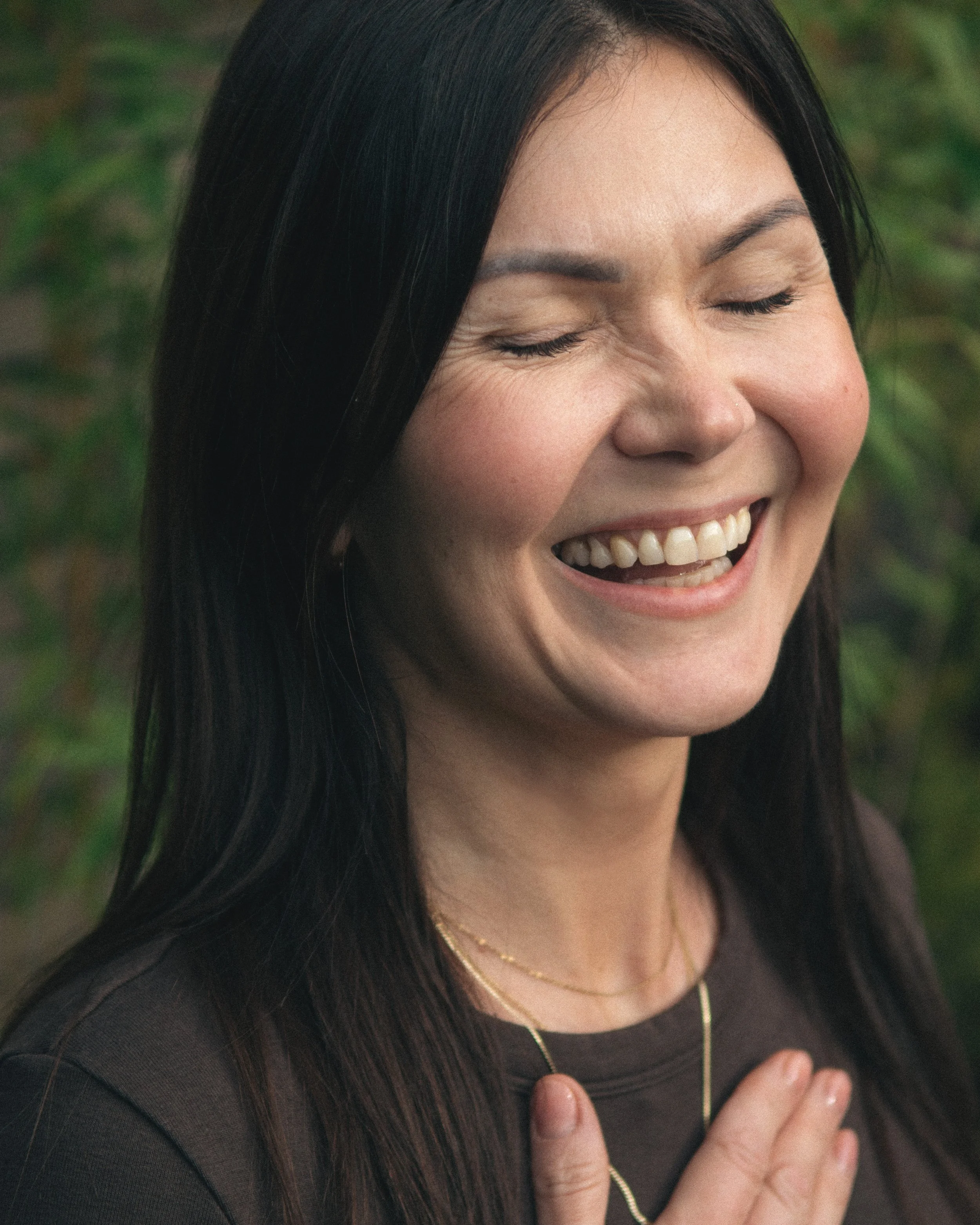 Close-up of a woman smiling with eyes closed, outdoors, with green foliage in the background.