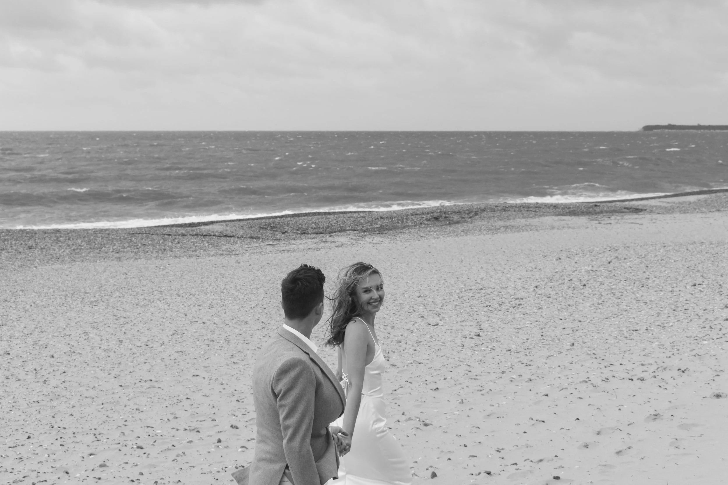 A black and white photo of a man and woman walking hand in hand on a sandy beach, with the ocean and cloudy sky in the background. The woman is smiling at the camera.