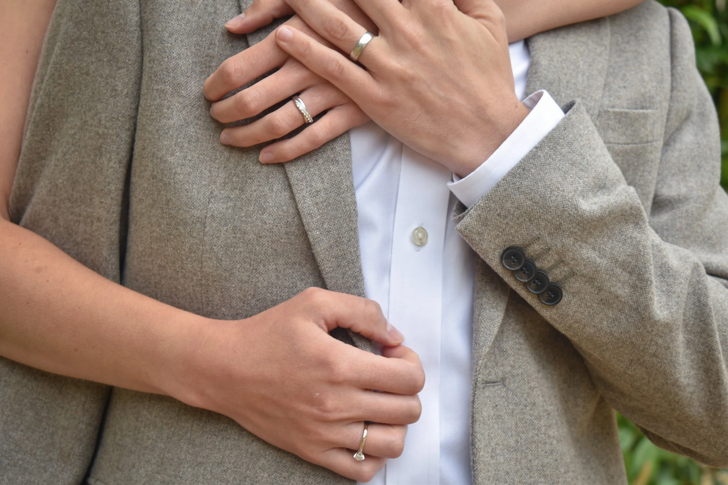 Close-up of a couple's hands showing wedding rings, with the woman hugging the man from behind, both dressed in formal attire.