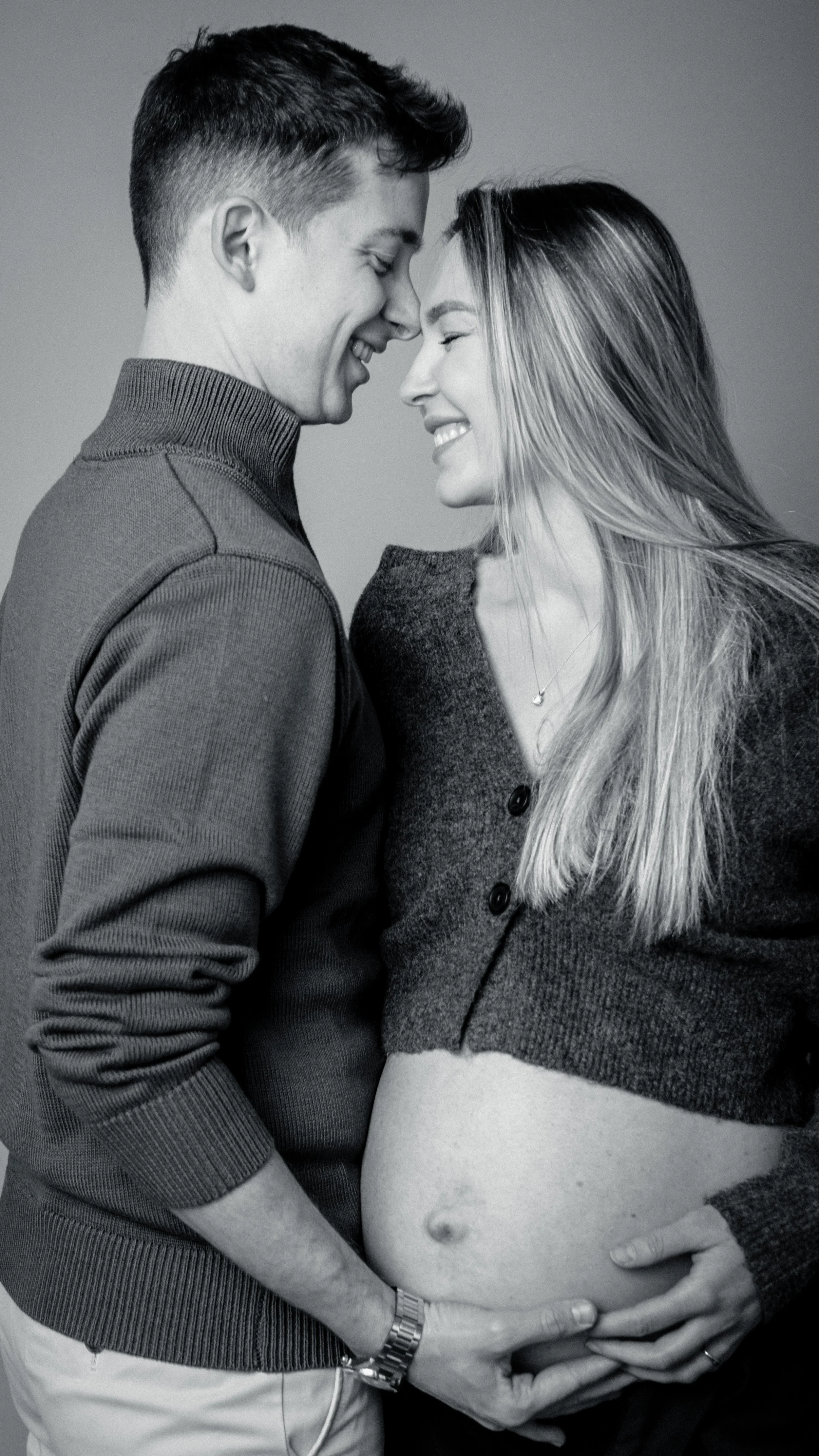 Black and white photo of a couple embracing, with the woman showing her pregnant belly and both touching it while smiling and touching foreheads.