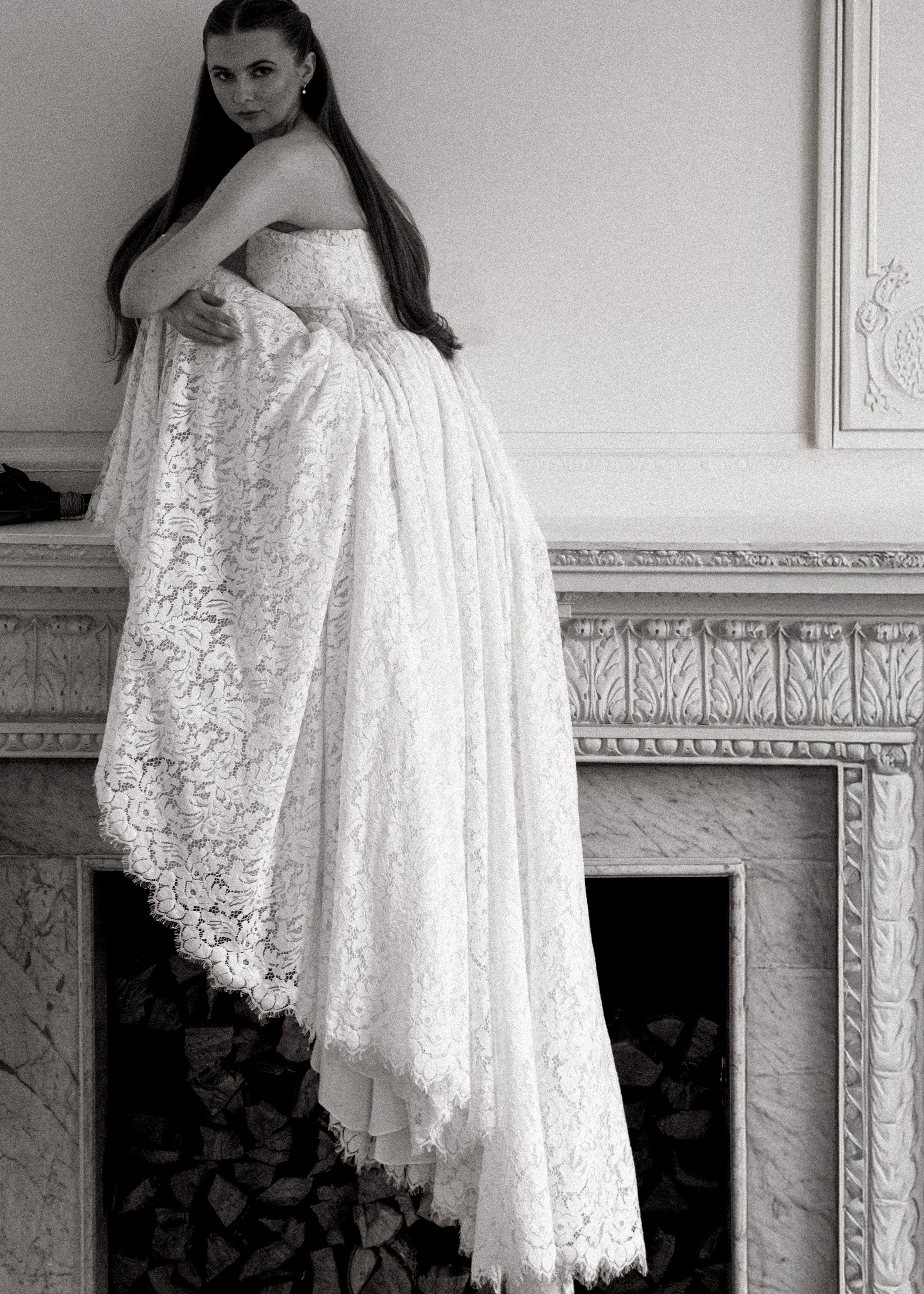 A woman in a strapless lace dress sitting on a fireplace mantle, looking at the camera, with her arms crossed over her knees. The fireplace has intricate carvings and a stack of firewood underneath.