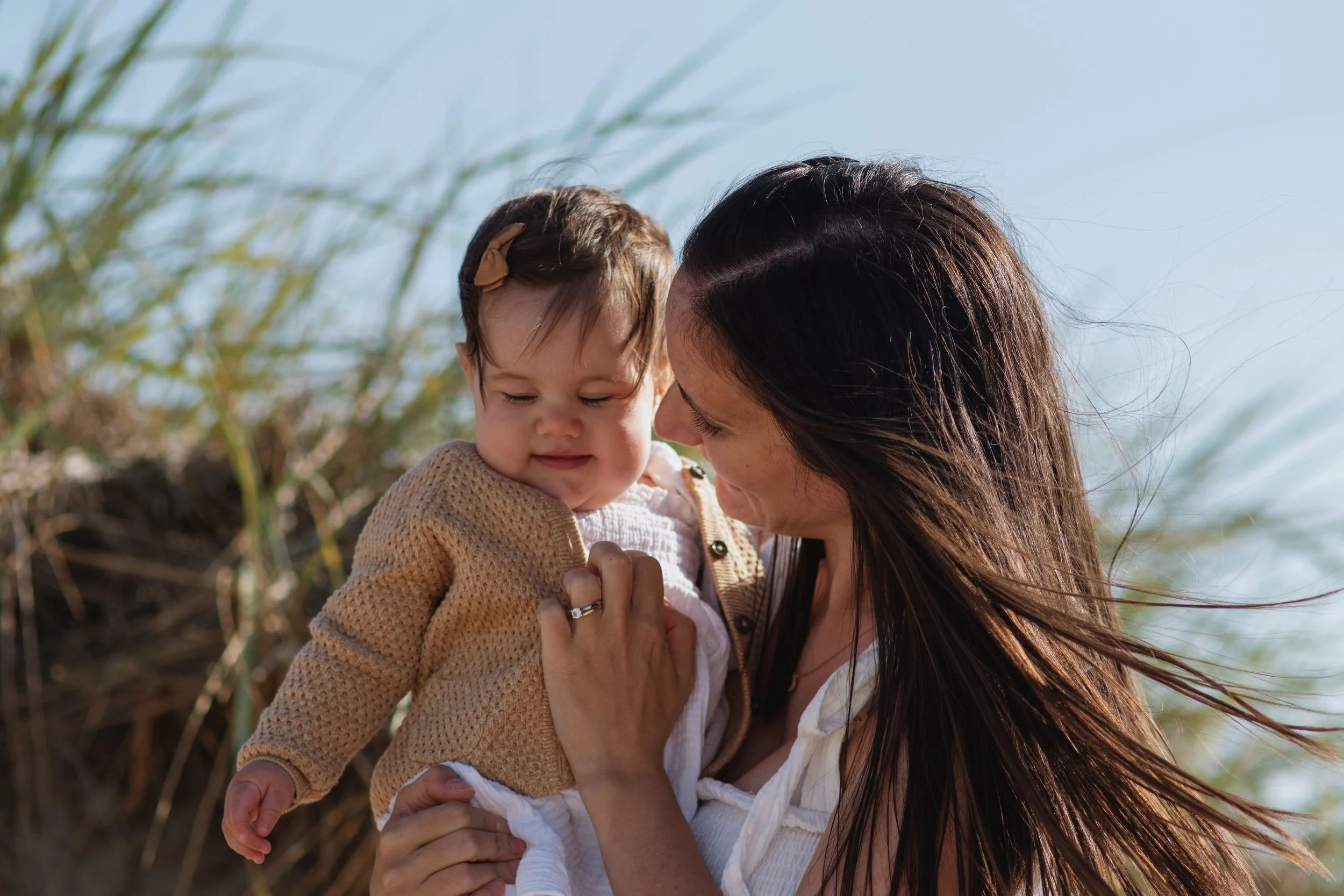 A woman holding a young child outdoors near tall grasses on a sunny day.