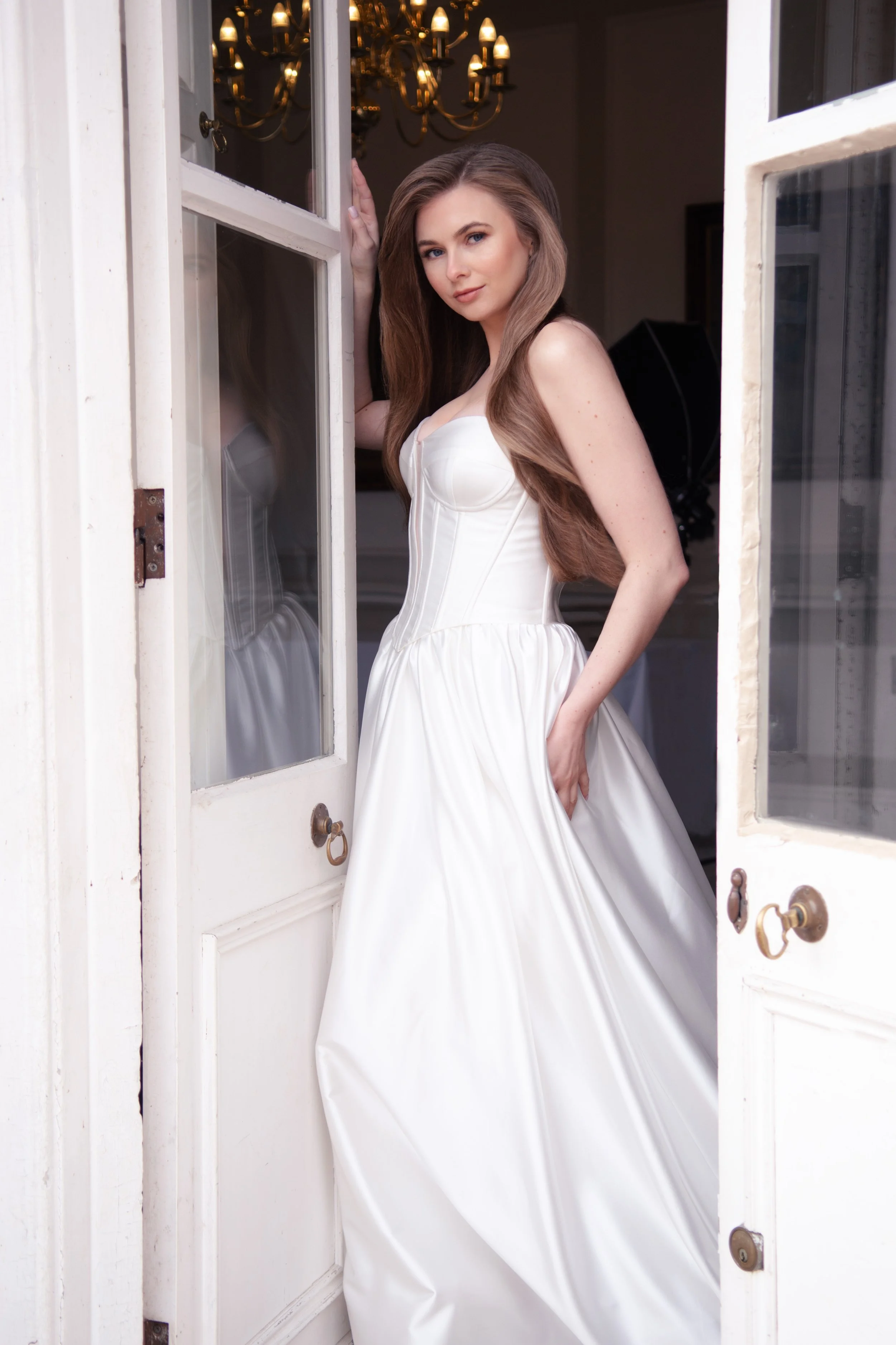 A woman in a strapless wedding gown standing in doorway, posing with one hand on the doorframe and the other in her dress.
