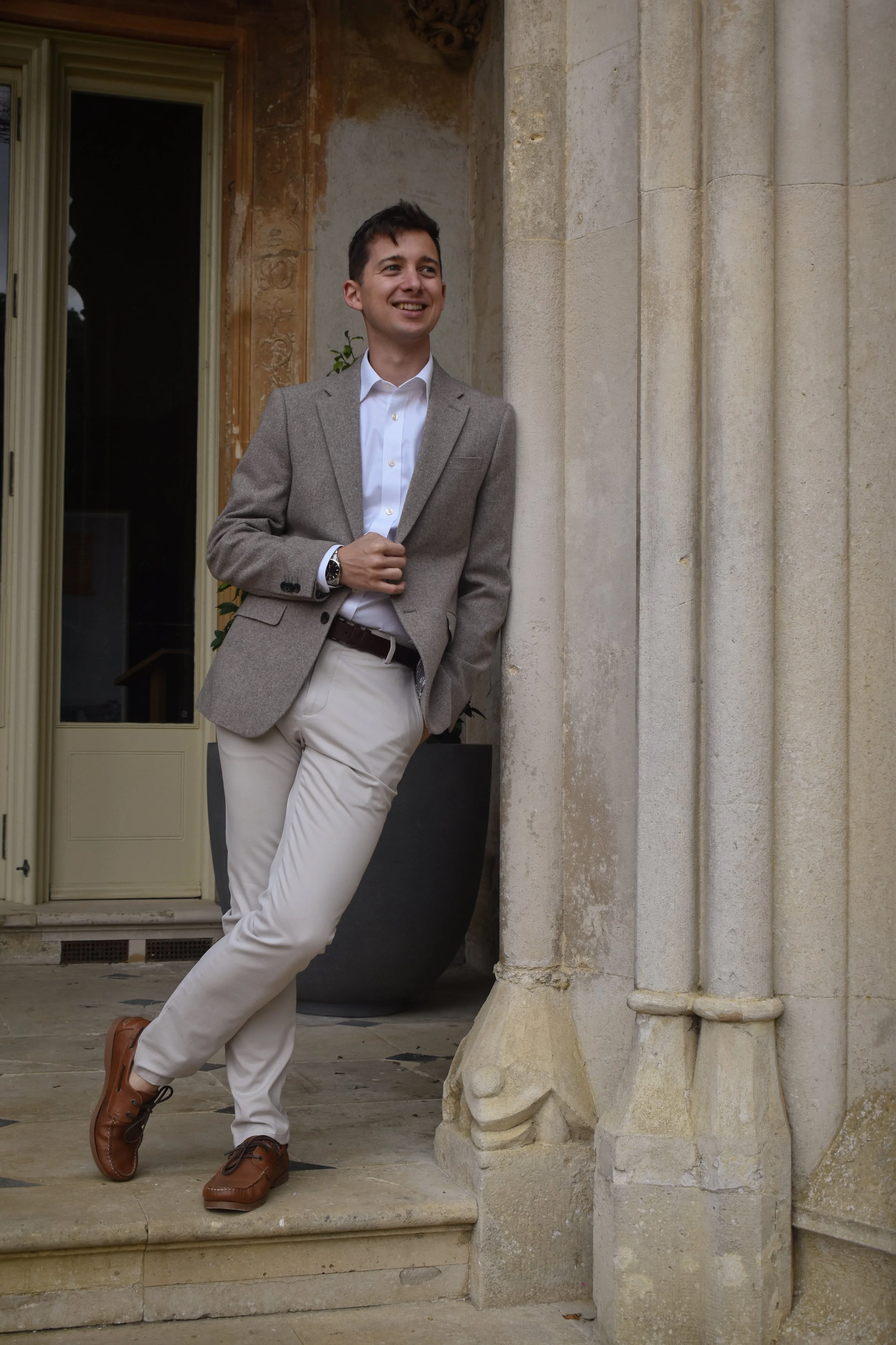 A young man in a gray blazer, white shirt, and beige pants, standing outdoors leaning against a stone building with columns, smiling and relaxed.