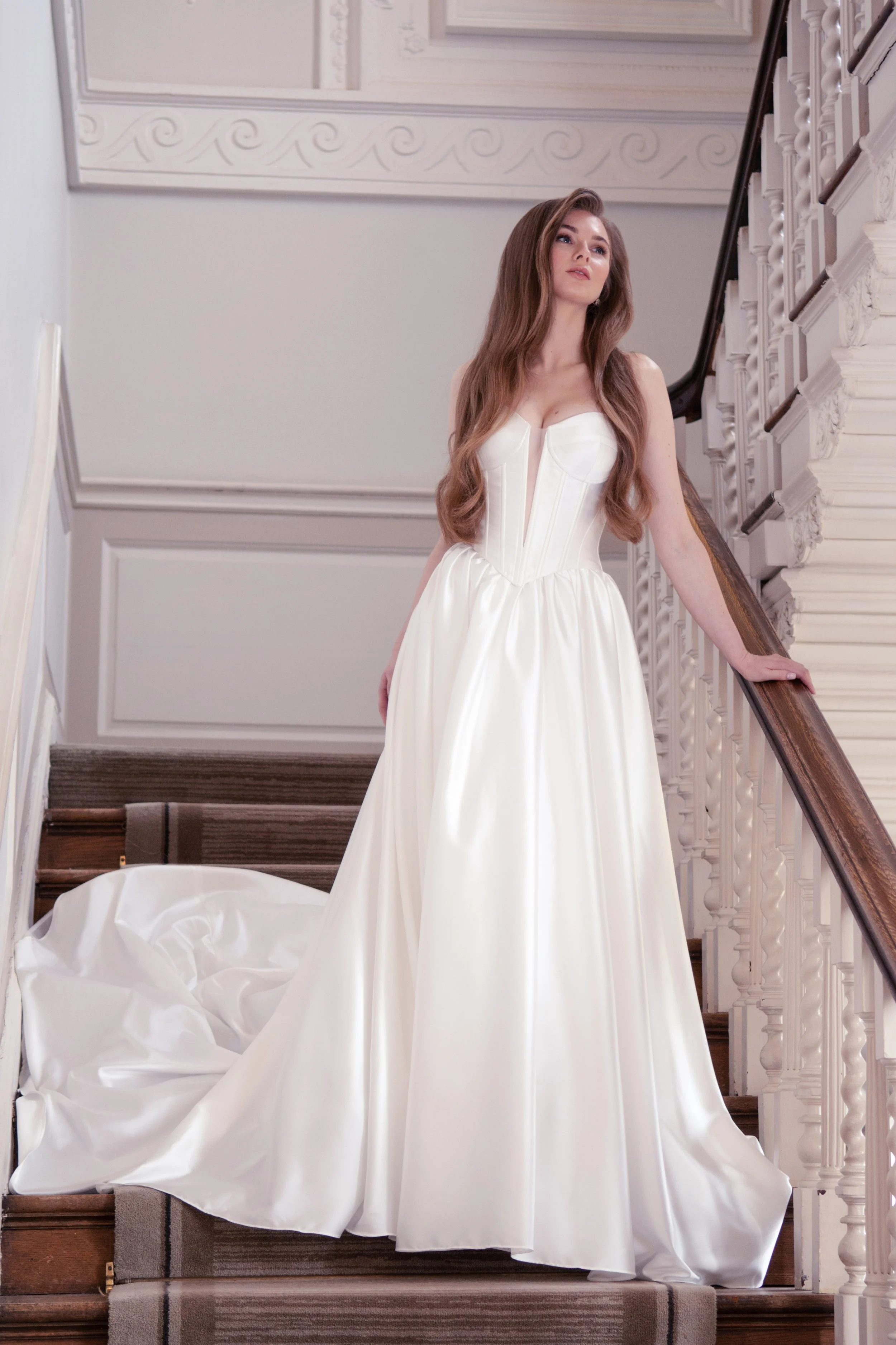 A woman in a white wedding dress standing on a staircase with a wooden banister and decorative wall paneling.