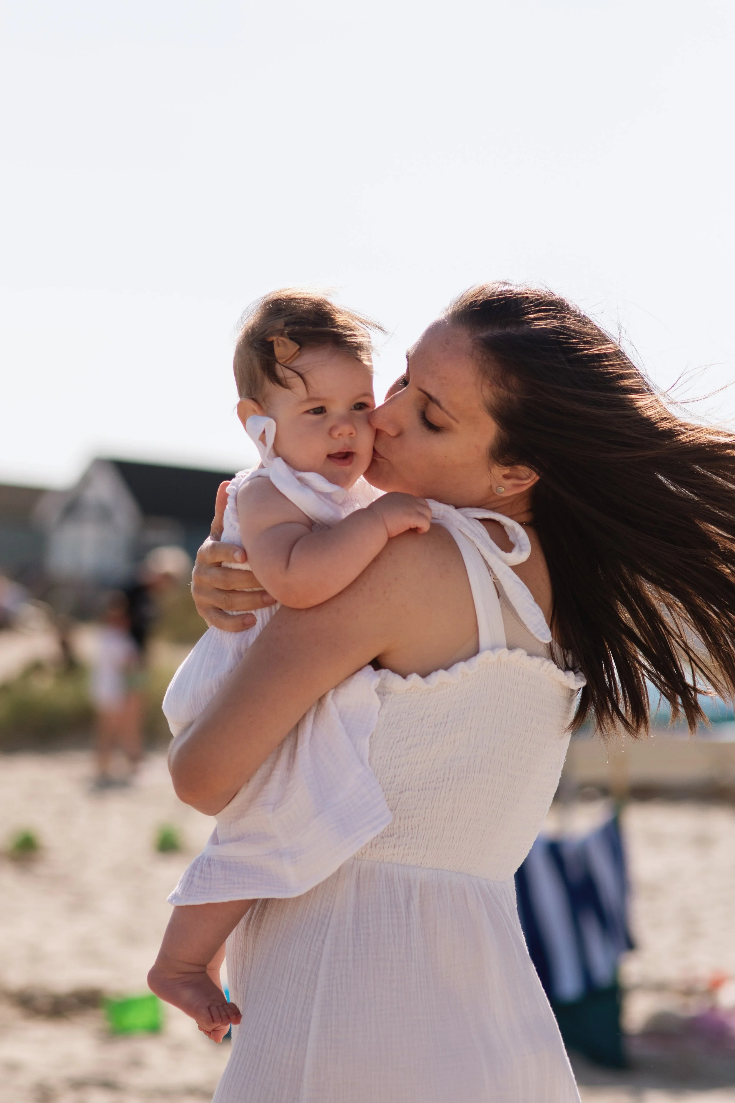 A woman holding a baby and kissing her cheek on a beach with sand, people, and houses in the background.