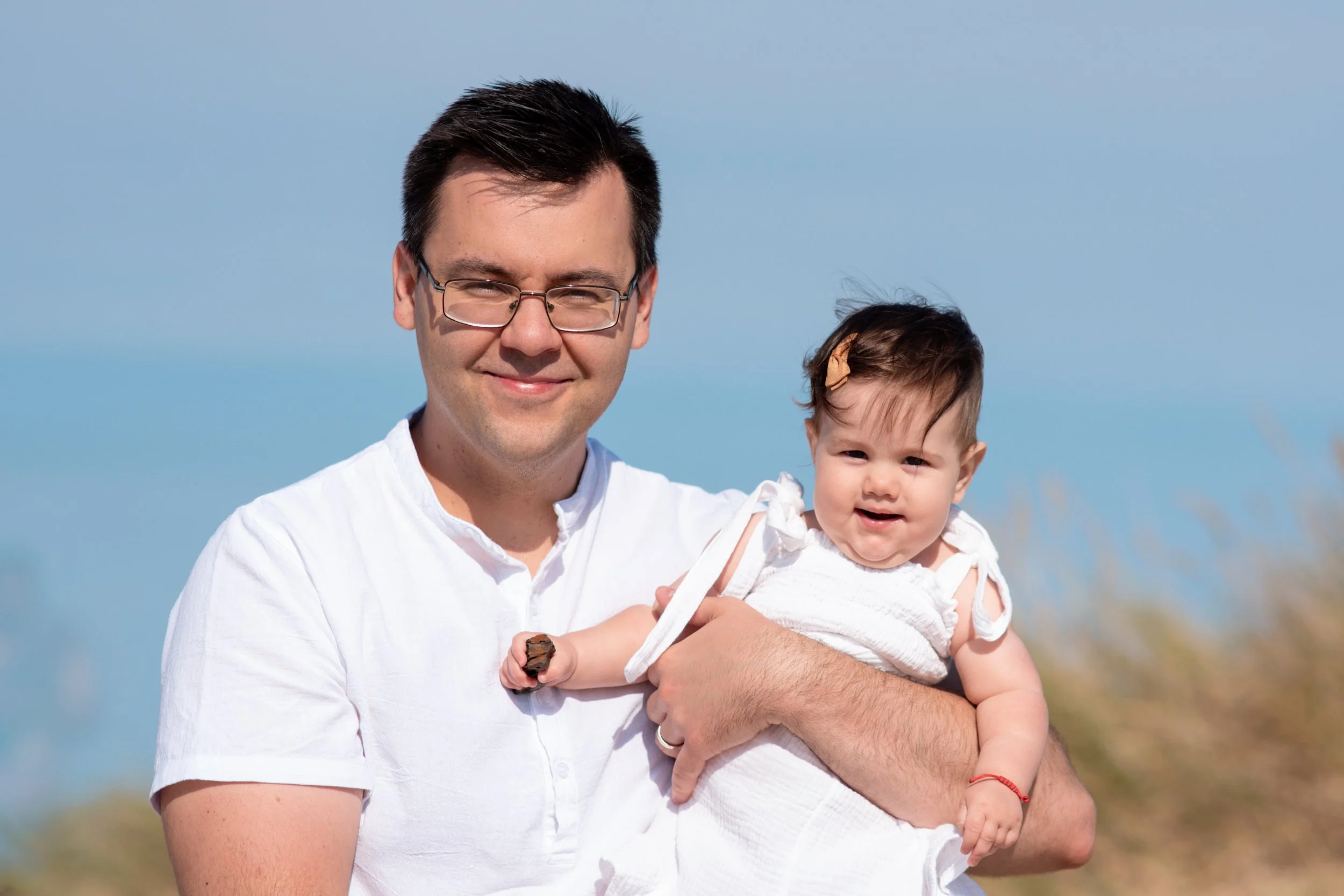 A man in a white shirt holding a young girl in white clothing outdoors with a blue sky background.