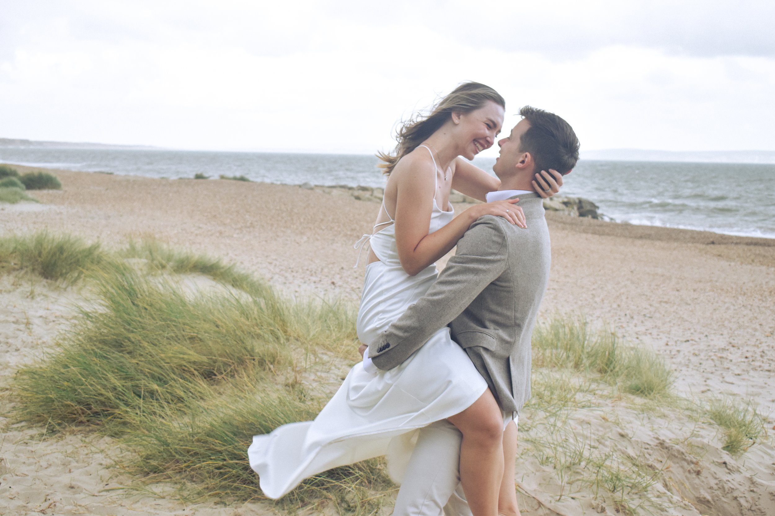 A couple on a beach, with the woman in a white dress and the man in a gray suit, embracing and smiling at each other.