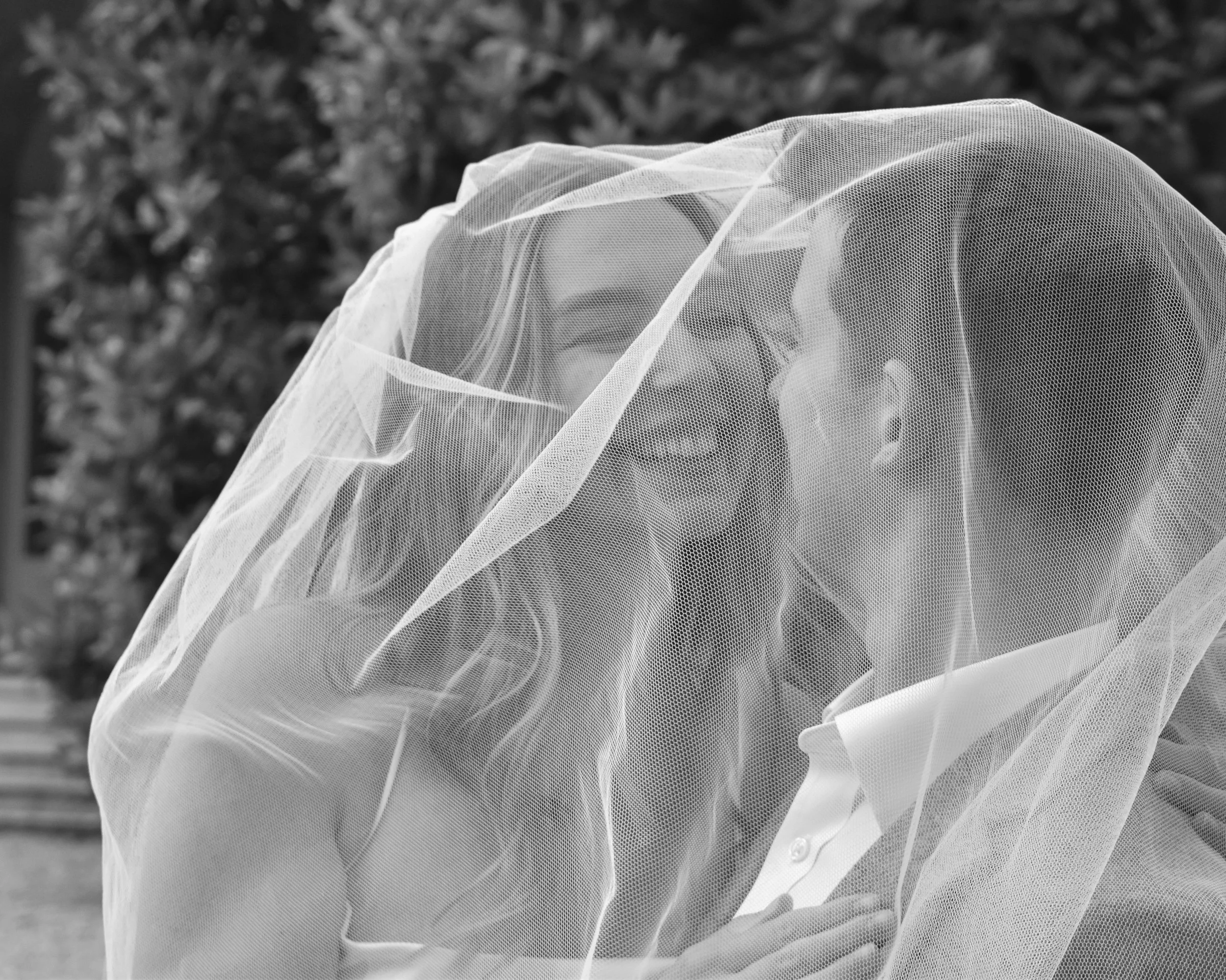 A woman and a man smiling and sharing a moment under a sheer veil outdoors, with trees in the background.