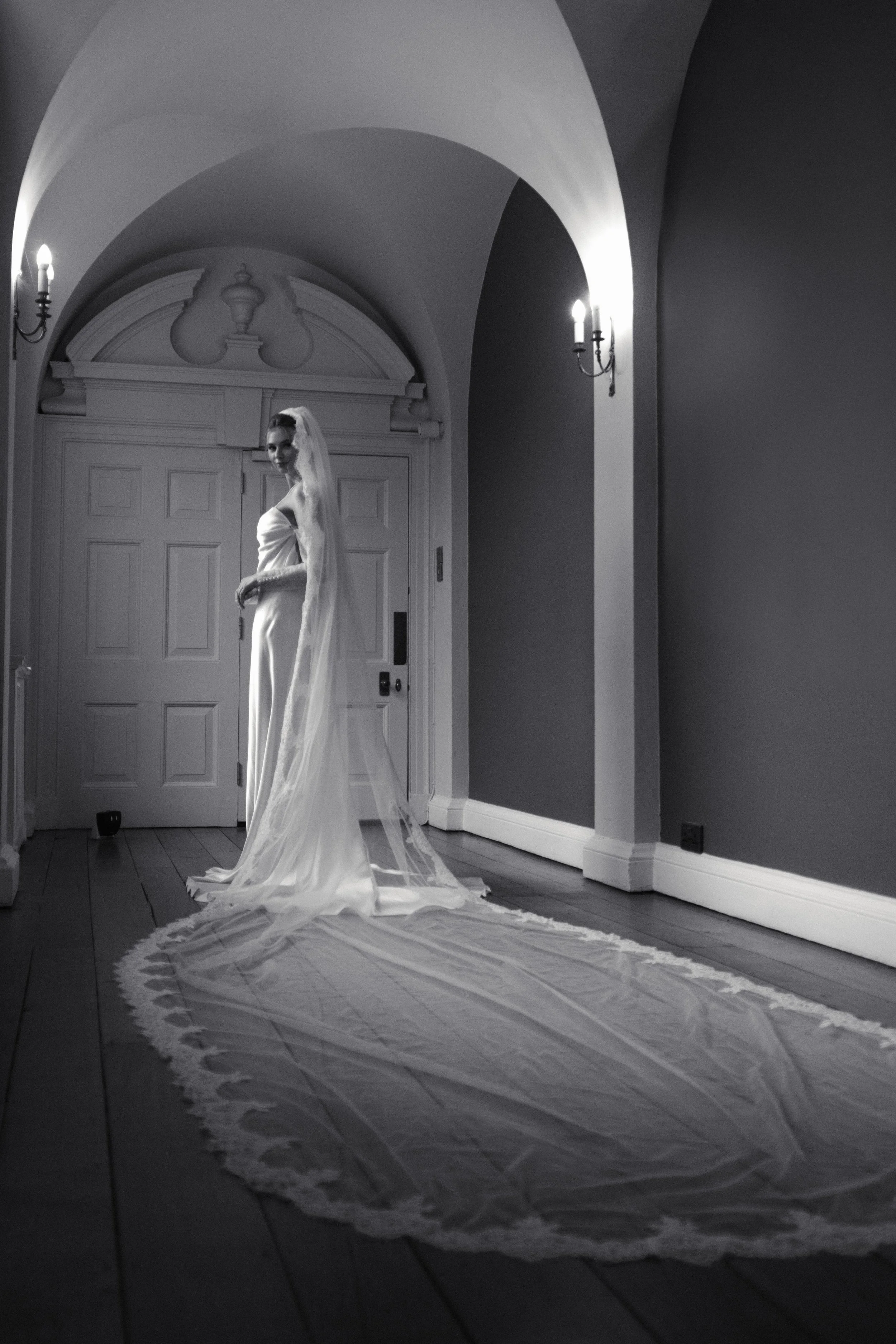 A bride in a wedding dress with a long veil standing in a hallway with wooden floors, arched ceilings, and wall sconces lighting, behind a closed door.