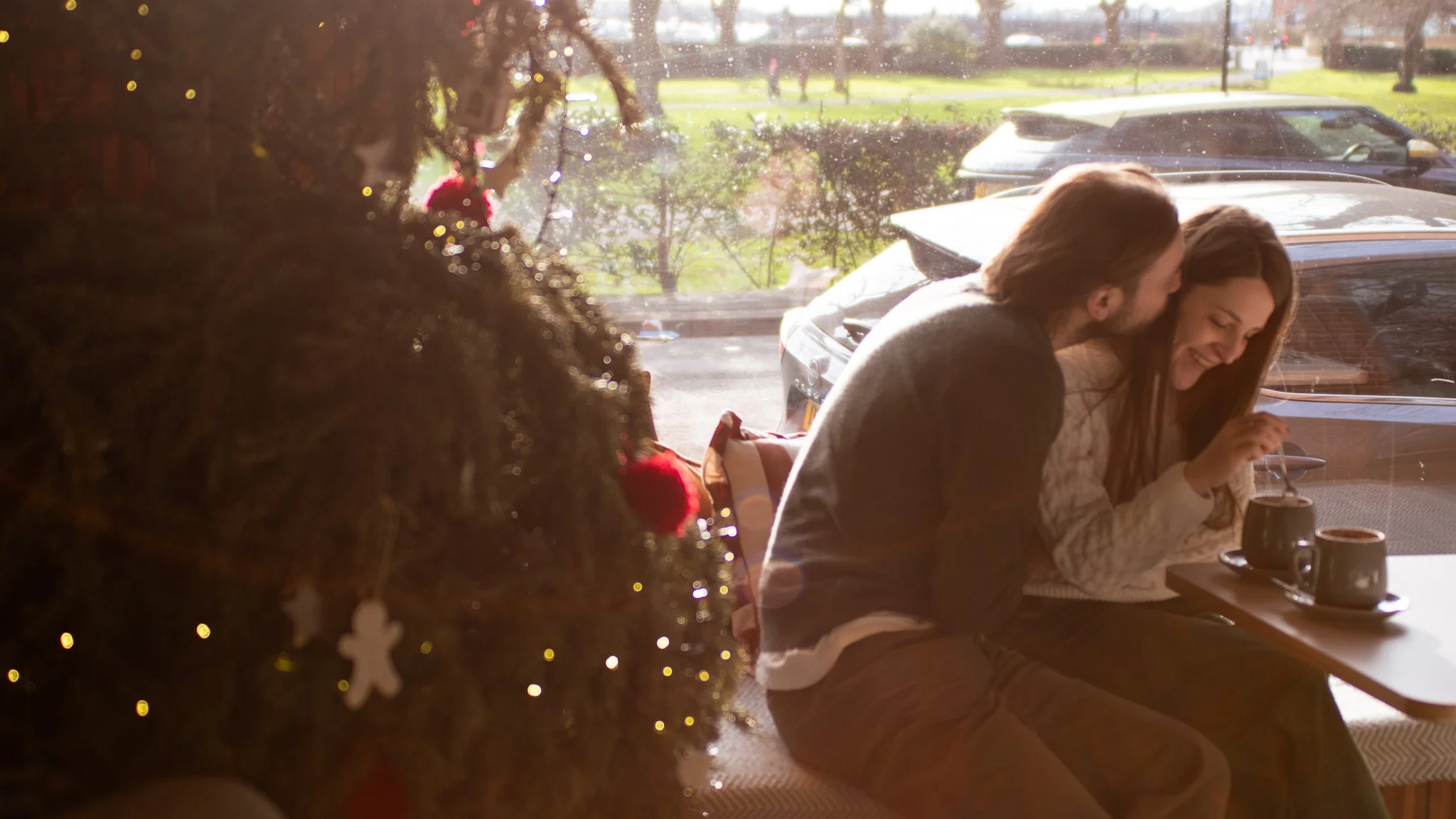 A couple sitting inside a cafe near a large window, sharing a tender moment with the man kissing the woman on the cheek while she smiles. There is Christmas decor on a nearby tree, and a parking lot with cars can be seen outside through the window.
