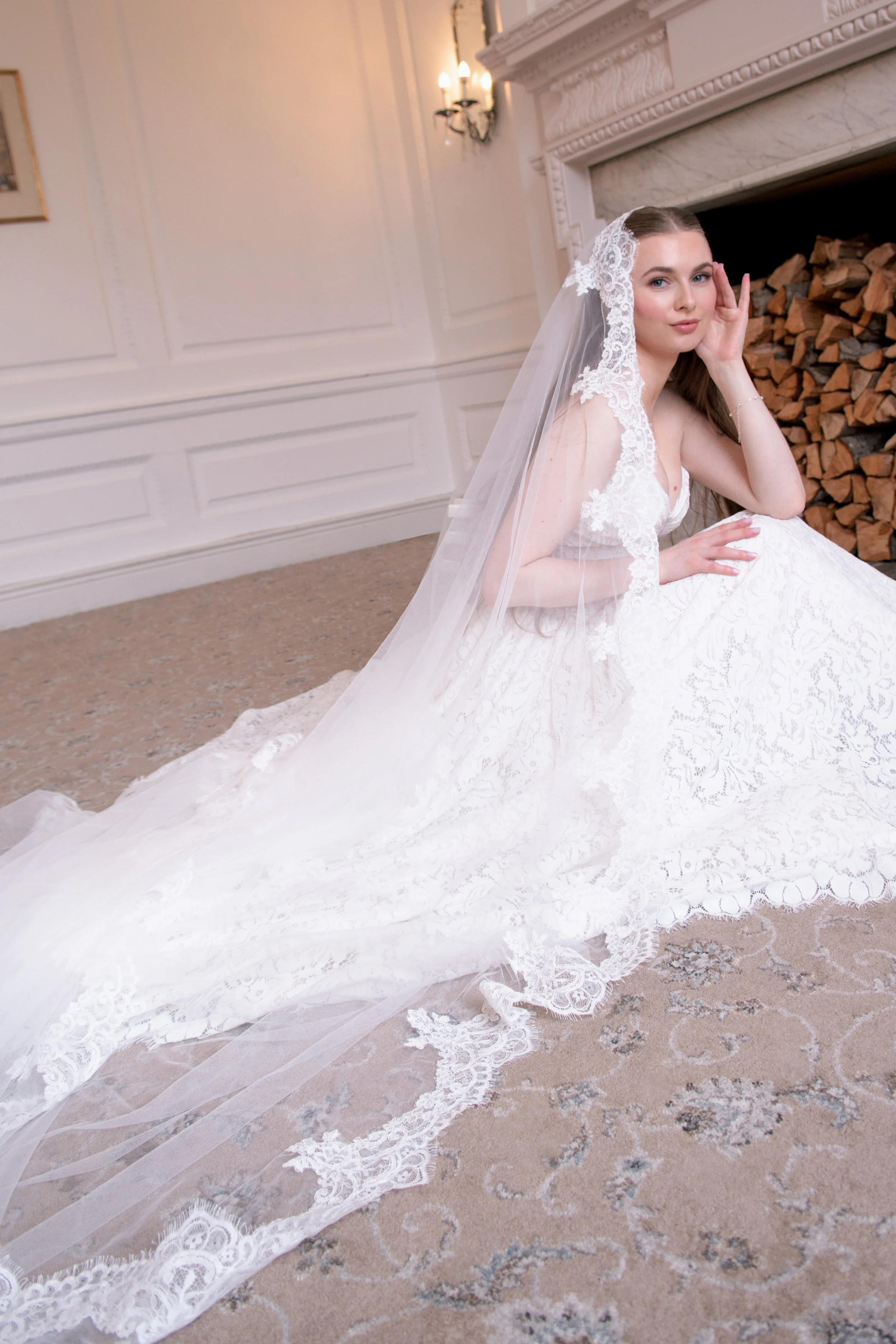 Bride sitting on the floor in a white wedding dress and veil, posing in front of a fireplace with wood logs. The room has white paneled walls and a chandelier.