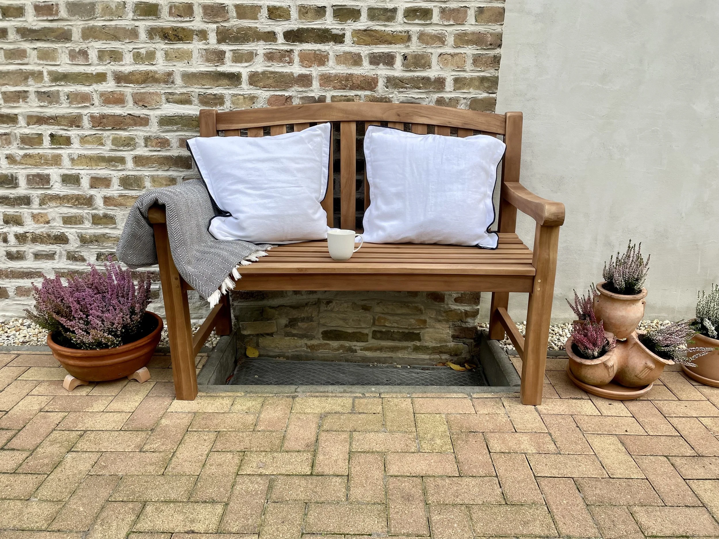 Ein Holzbank mit zwei weißen Kissen und einer Tasse steht an einer Steinmauer. Aktivitäten im Garten, umgeben von Blumentöpfen mit Heidestrauchpflanzen.