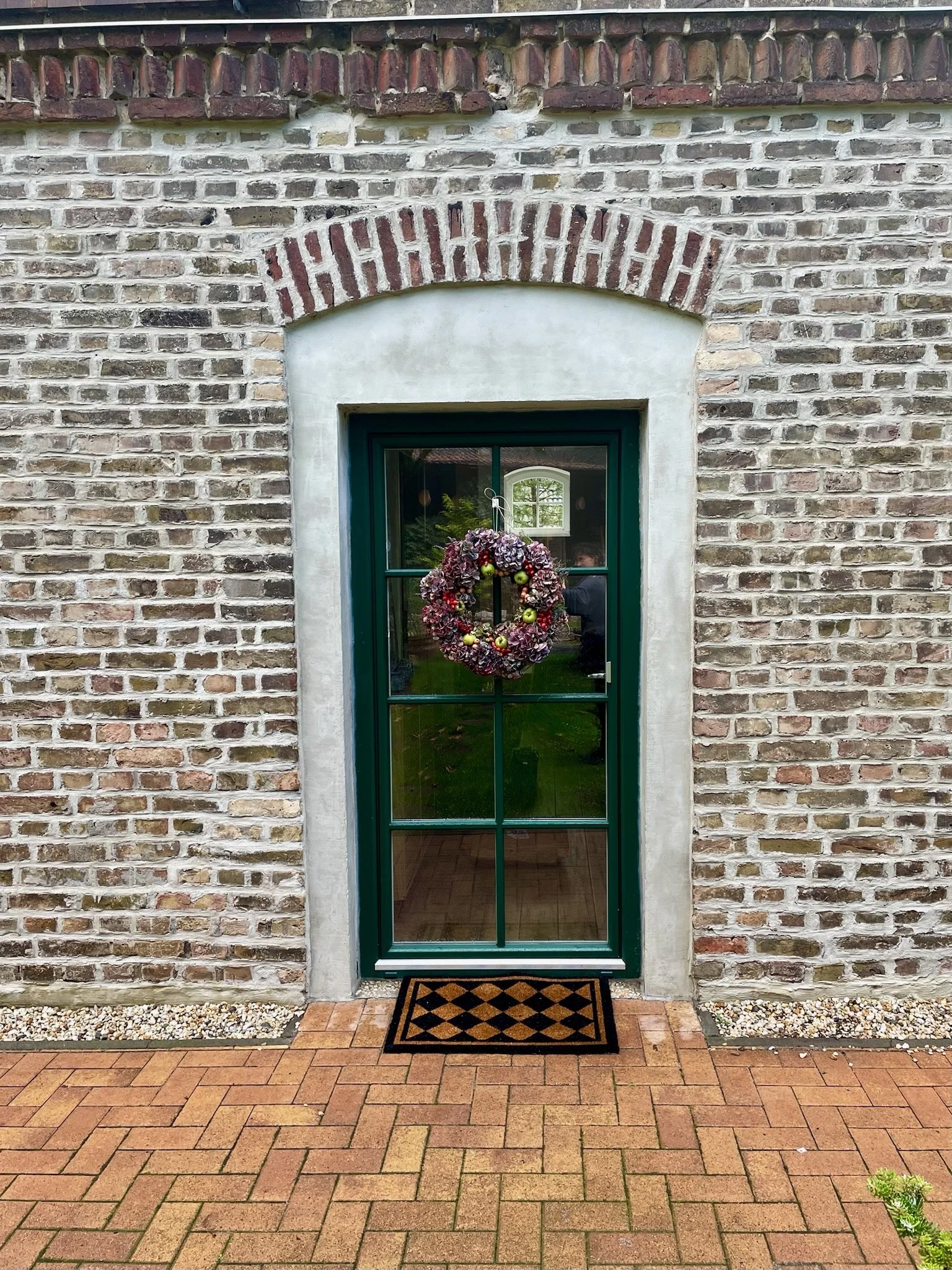 Eine Haustür mit grünem Rahmen und einem weihnachtlichen Kranz darüber, umgeben von einer Backsteinwand und einem gepflasterten Boden.