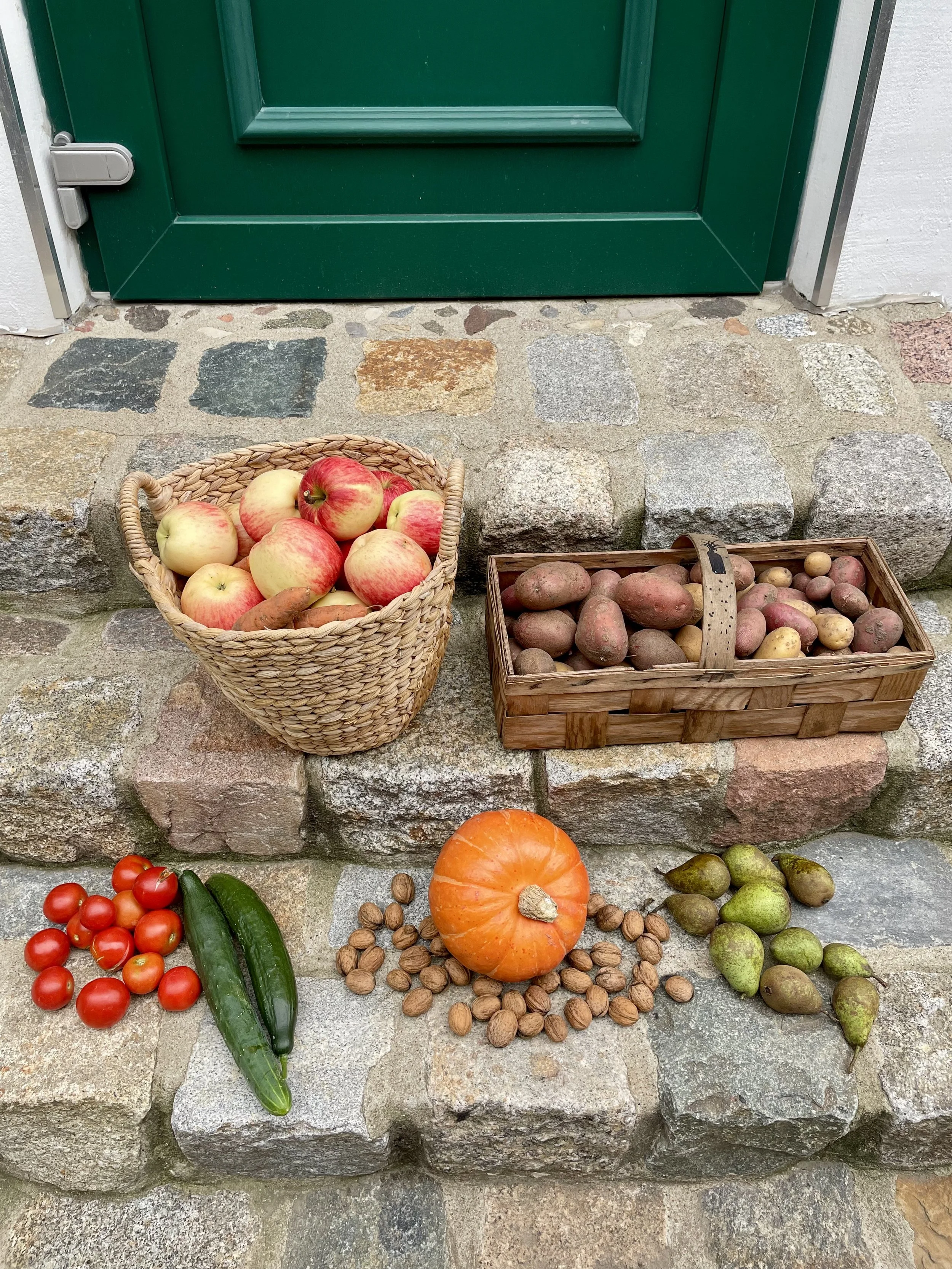 Bunte Früchte und Gemüse auf Steinstufen vor einer grünen Tür, inklusive Äpfel in einem Körbchen, Kartoffeln in einem Holzkistchen, Tomaten, Gurken, Kürbis, Walnüsse und Birnen.