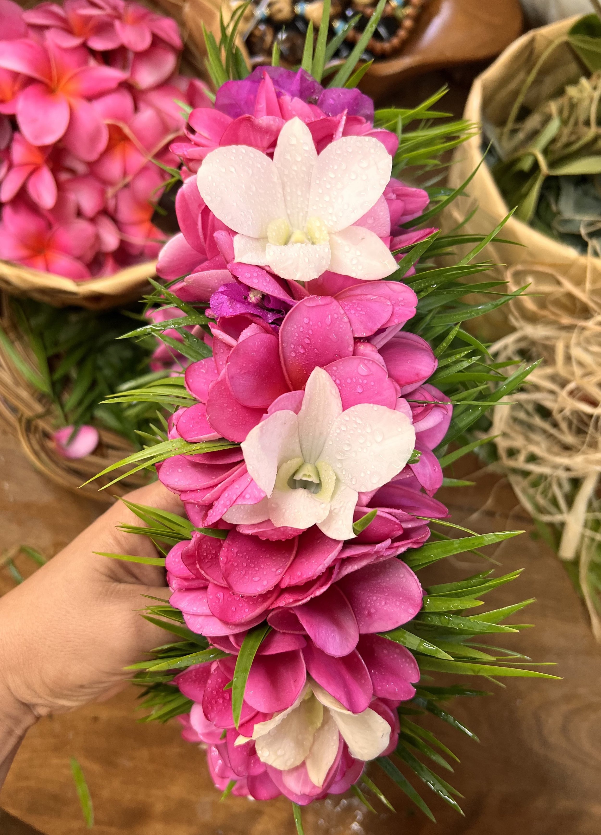A hand holding a vertical arrangement of pink and white flowers with water droplets, surrounded by green leaves and wicker baskets in the background.