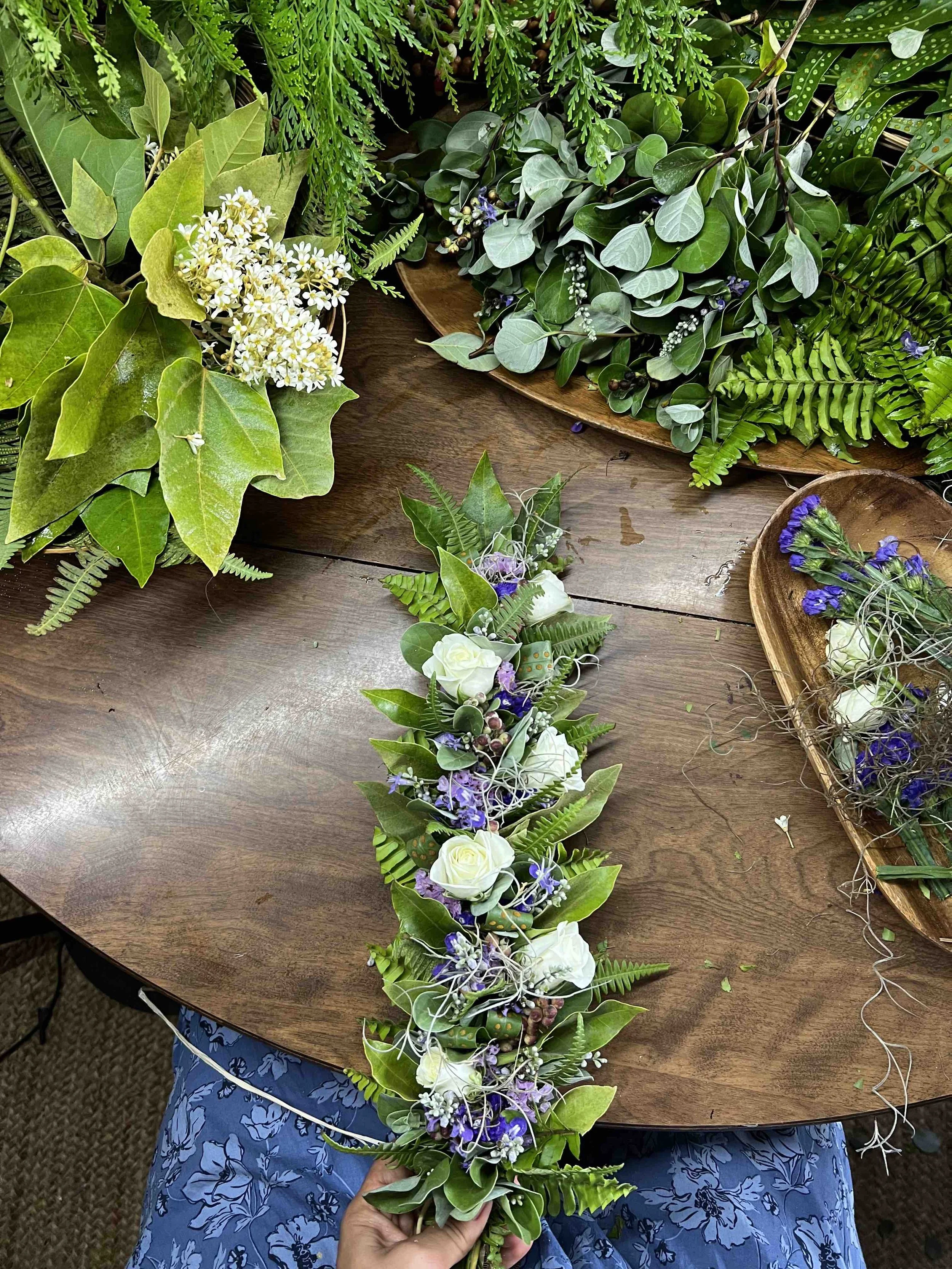 Floral arrangement on a wooden table with green foliage, white roses, purple flowers, and various leaves.