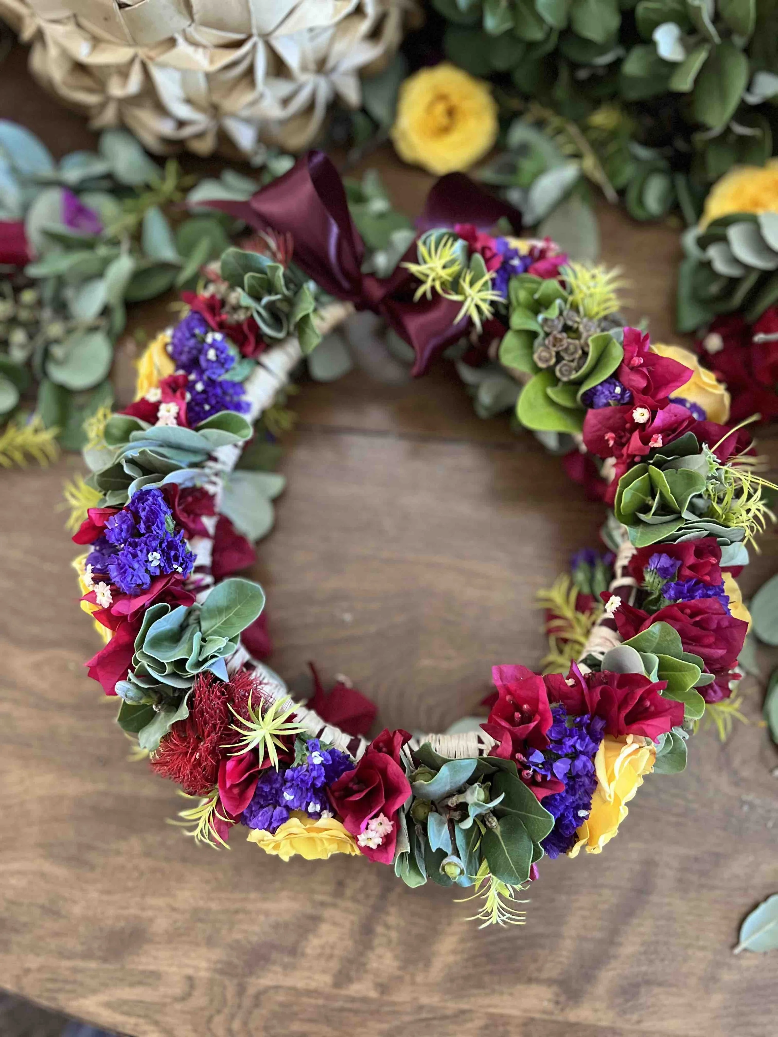 A colorful floral wreath made of various flowers and green leaves, with a dark red ribbon bow, resting on a wooden surface.