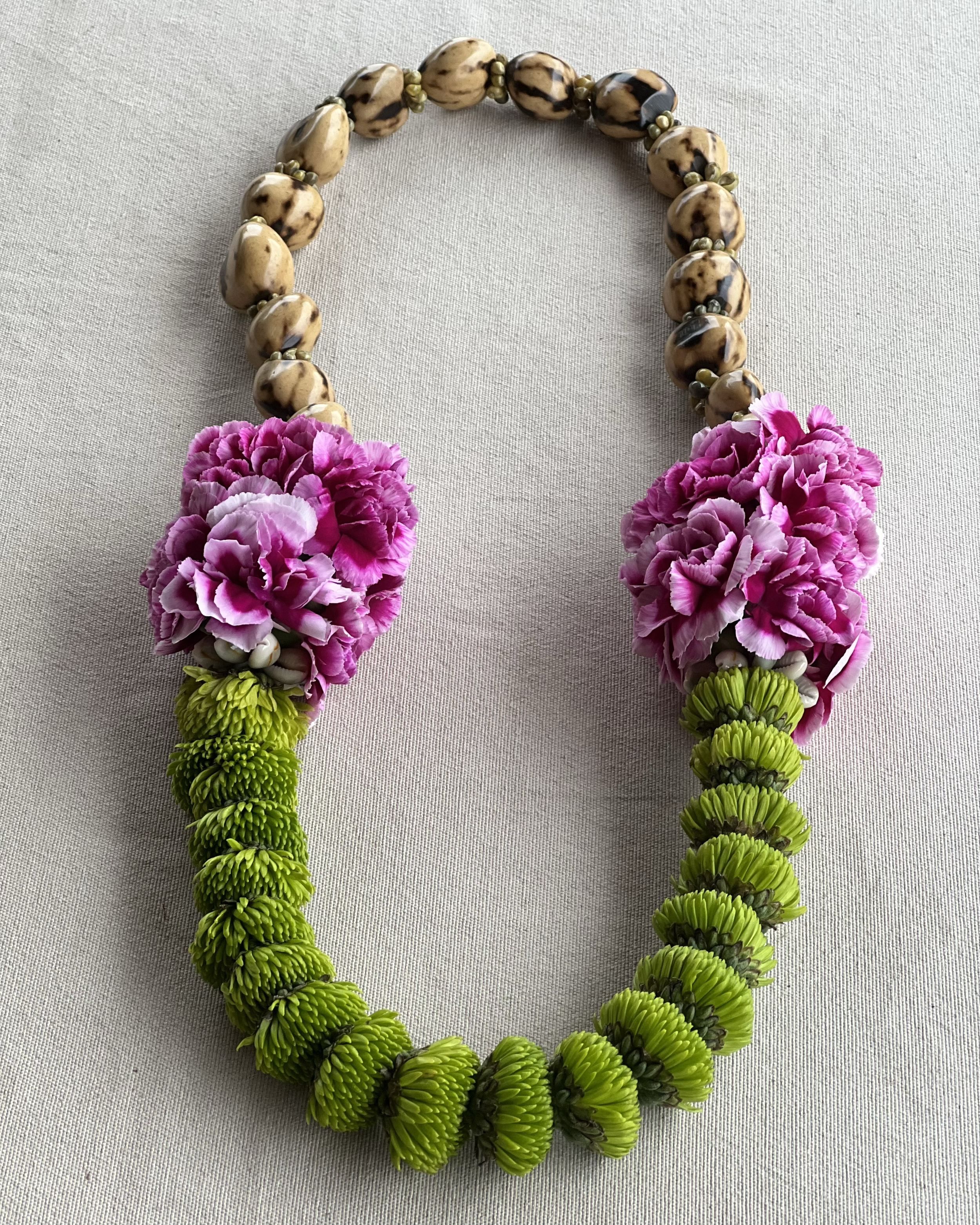 A floral necklace made of brown and black speckled beads, pink carnations, and green chrysanthemums, laid on a light gray fabric background.